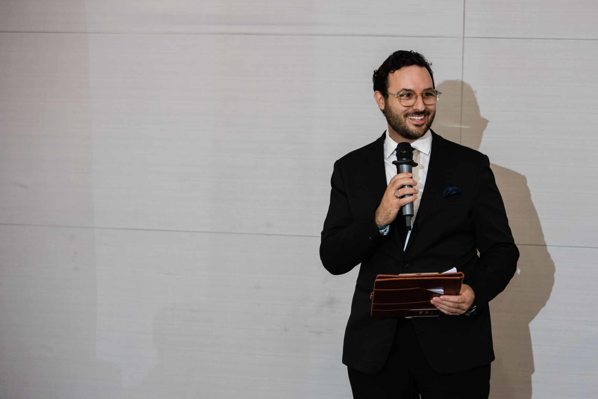 Well-dressed wedding emcee holding a microphone and folder while speaking indoors.