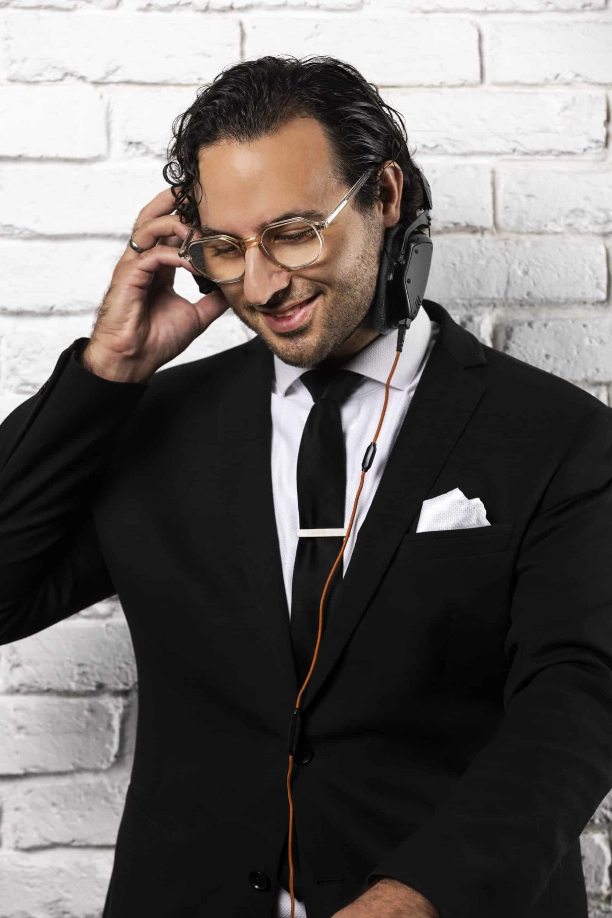 Smiling wedding DJ in a black suit and headphones standing against a white brick wall.