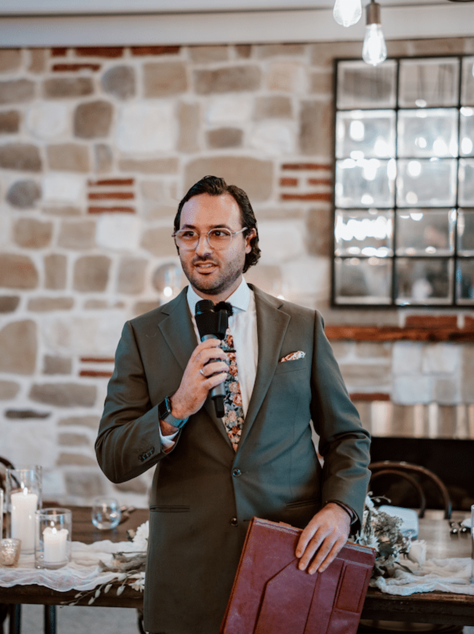 Well-dressed wedding officiant holding a microphone and folder while speaking indoors by a stone wall.