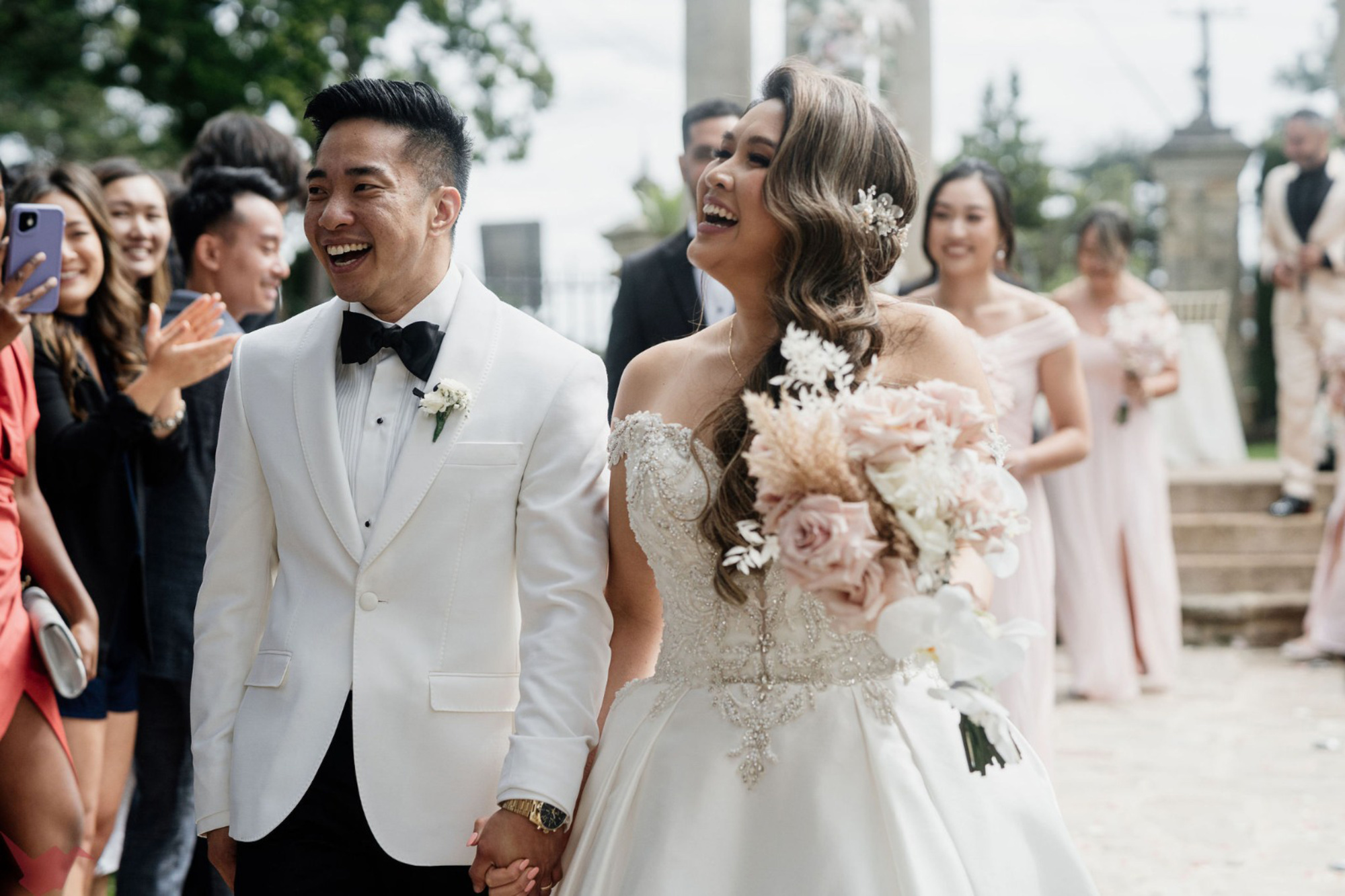 Smiling bride and groom walk hand in hand down the aisle outdoors surrounded by cheering guests.