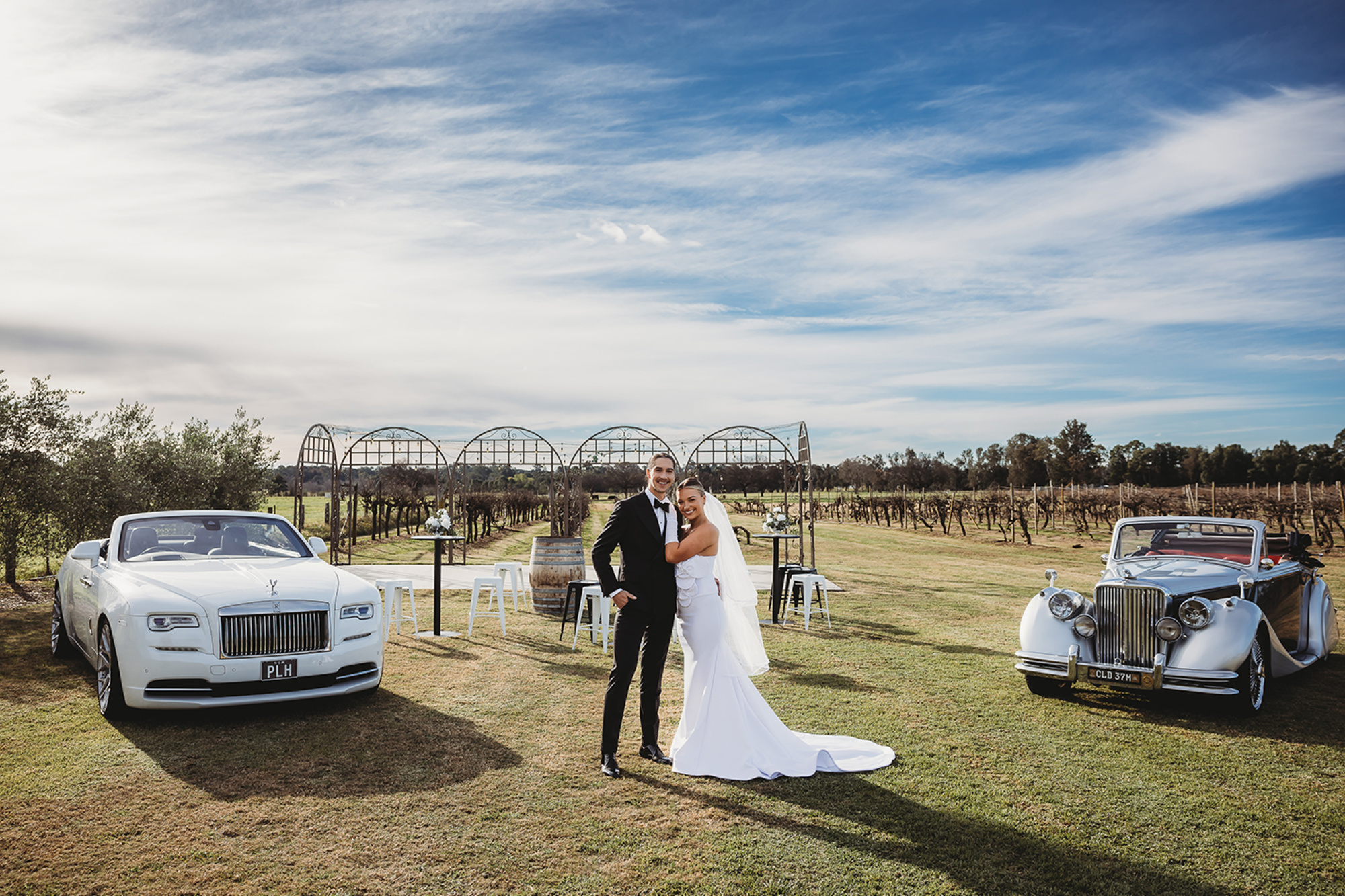 Bride and groom pose between two luxury convertibles in a sunny vineyard wedding setting.