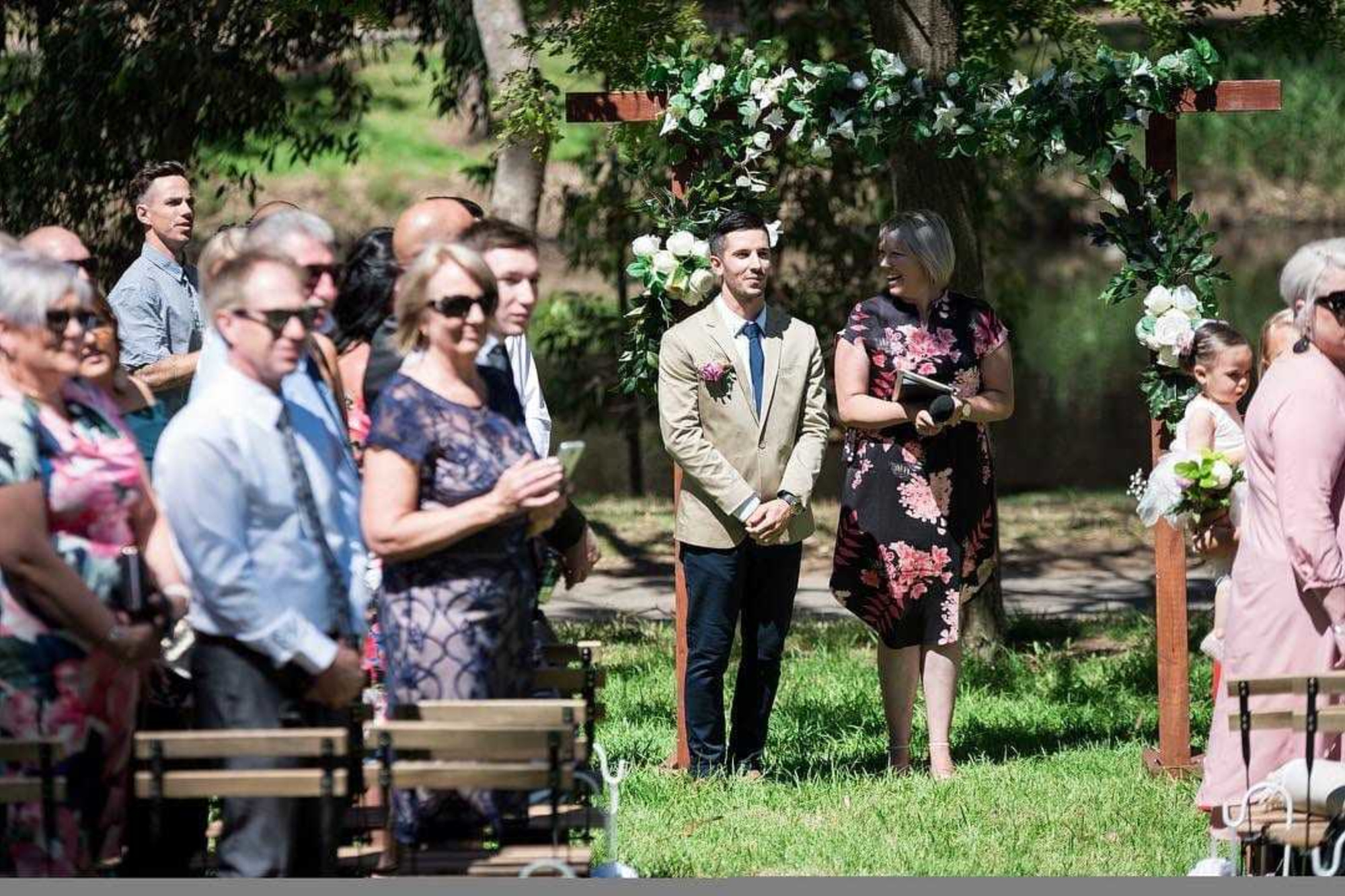 Outdoor wedding ceremony with the groom waiting under a floral arch as guests stand around.