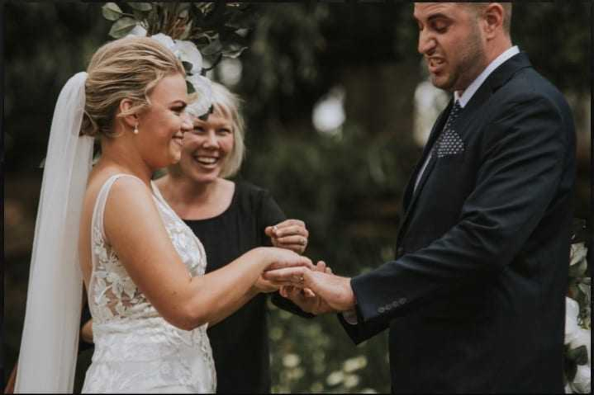 Bride and groom exchange rings during an outdoor wedding ceremony with the officiant smiling beside them.