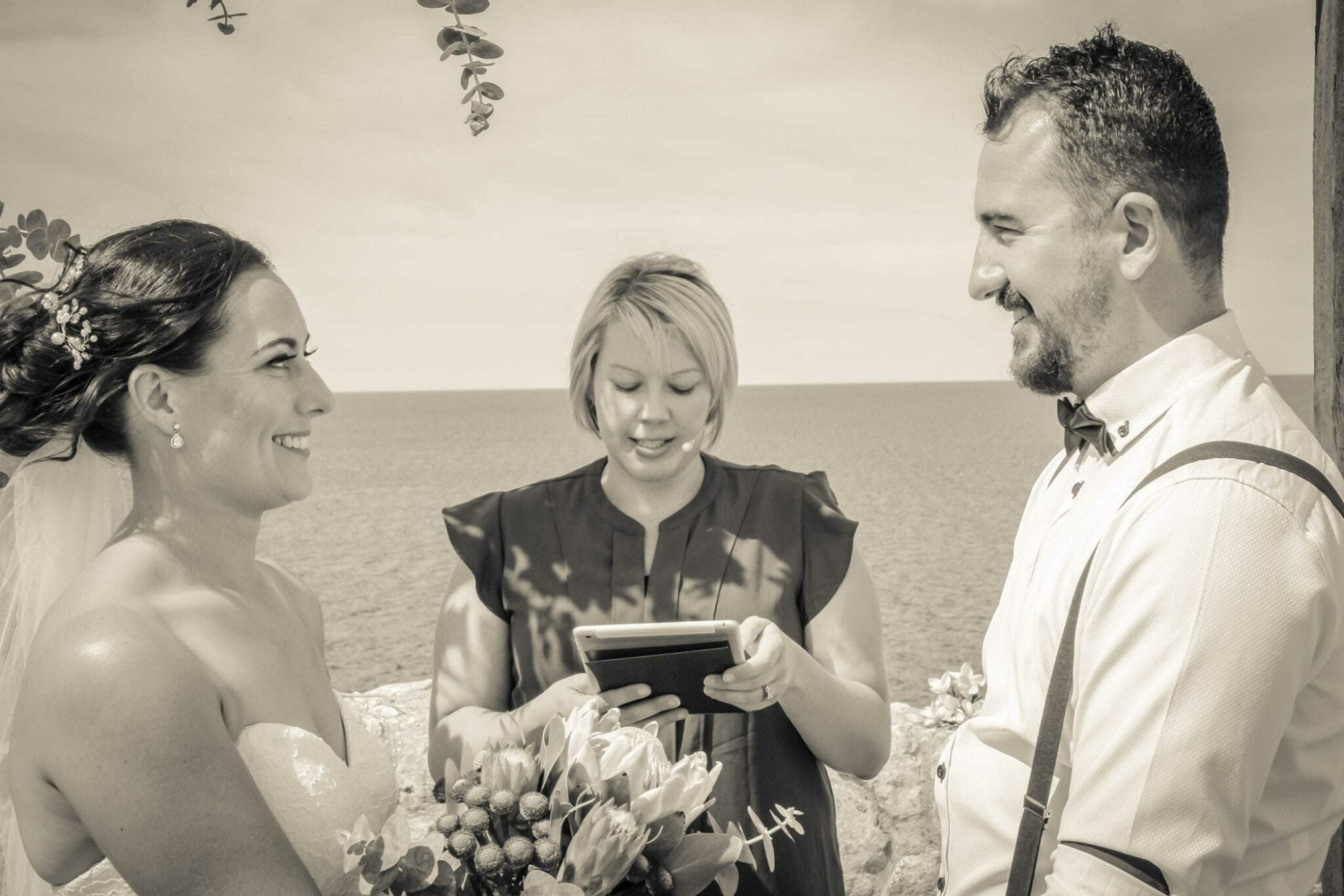 Bride and groom smiling at each other during an outdoor seaside ceremony led by an officiant.