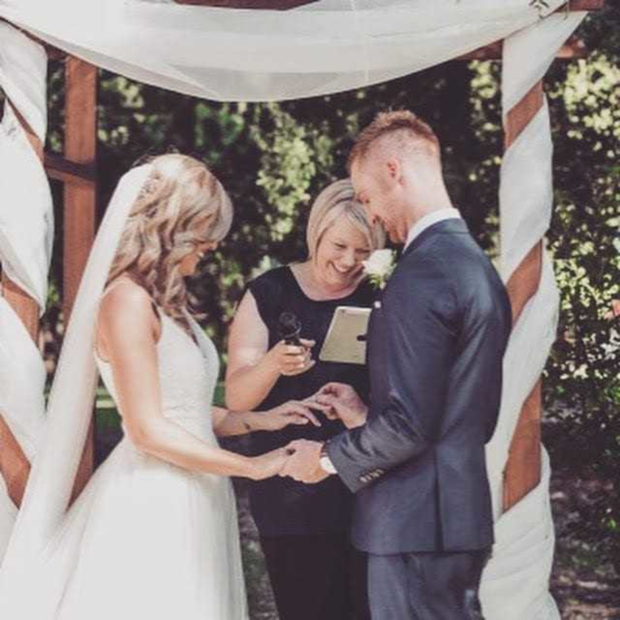 An outdoor wedding ceremony where a couple exchanges rings under a draped wooden arch with an officiant.
