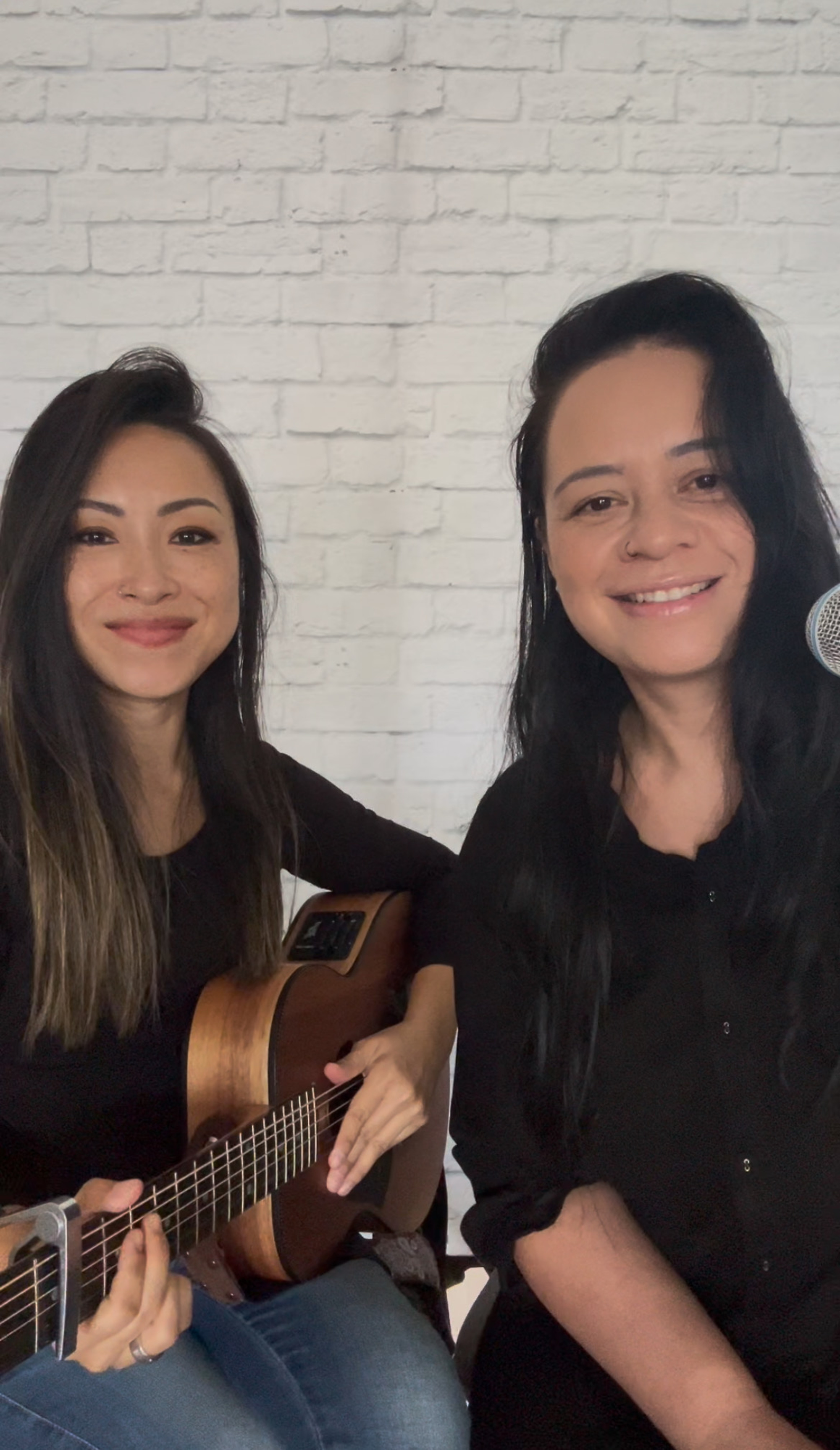 Two smiling musicians with a guitar and microphone posing against a white brick wall.