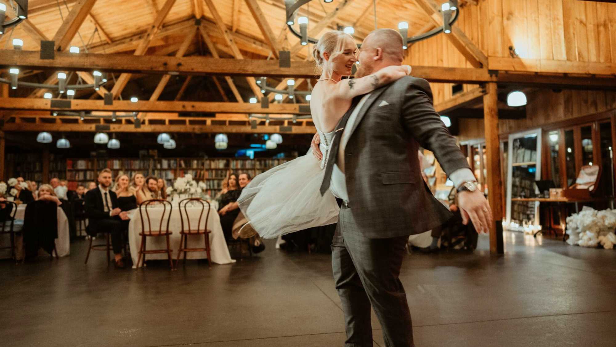Bride and groom share a joyful first dance in a warm wooden barn-style wedding venue surrounded by seated guests.