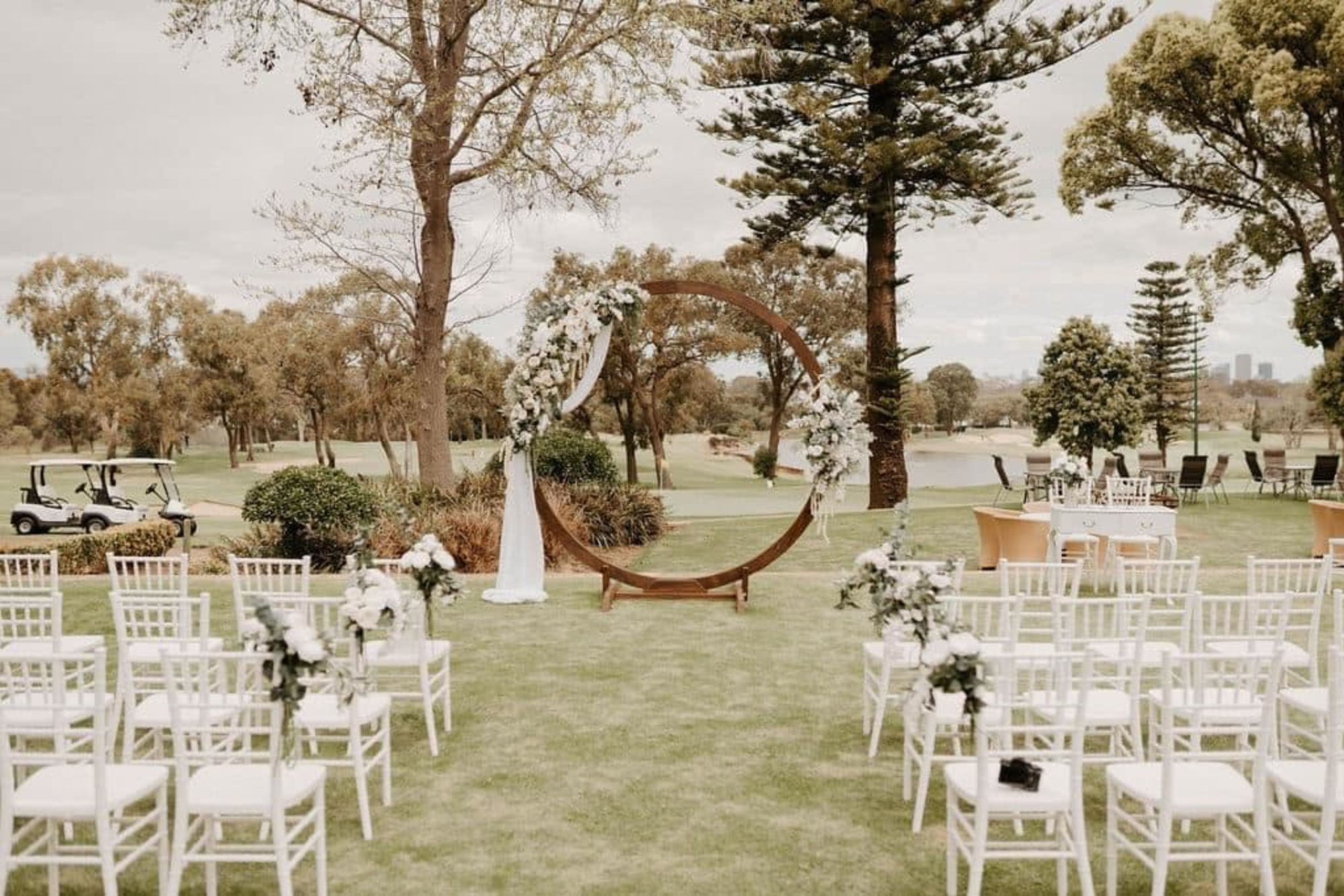 Outdoor wedding ceremony setup on a golf course with a circular floral arch and rows of white chairs.