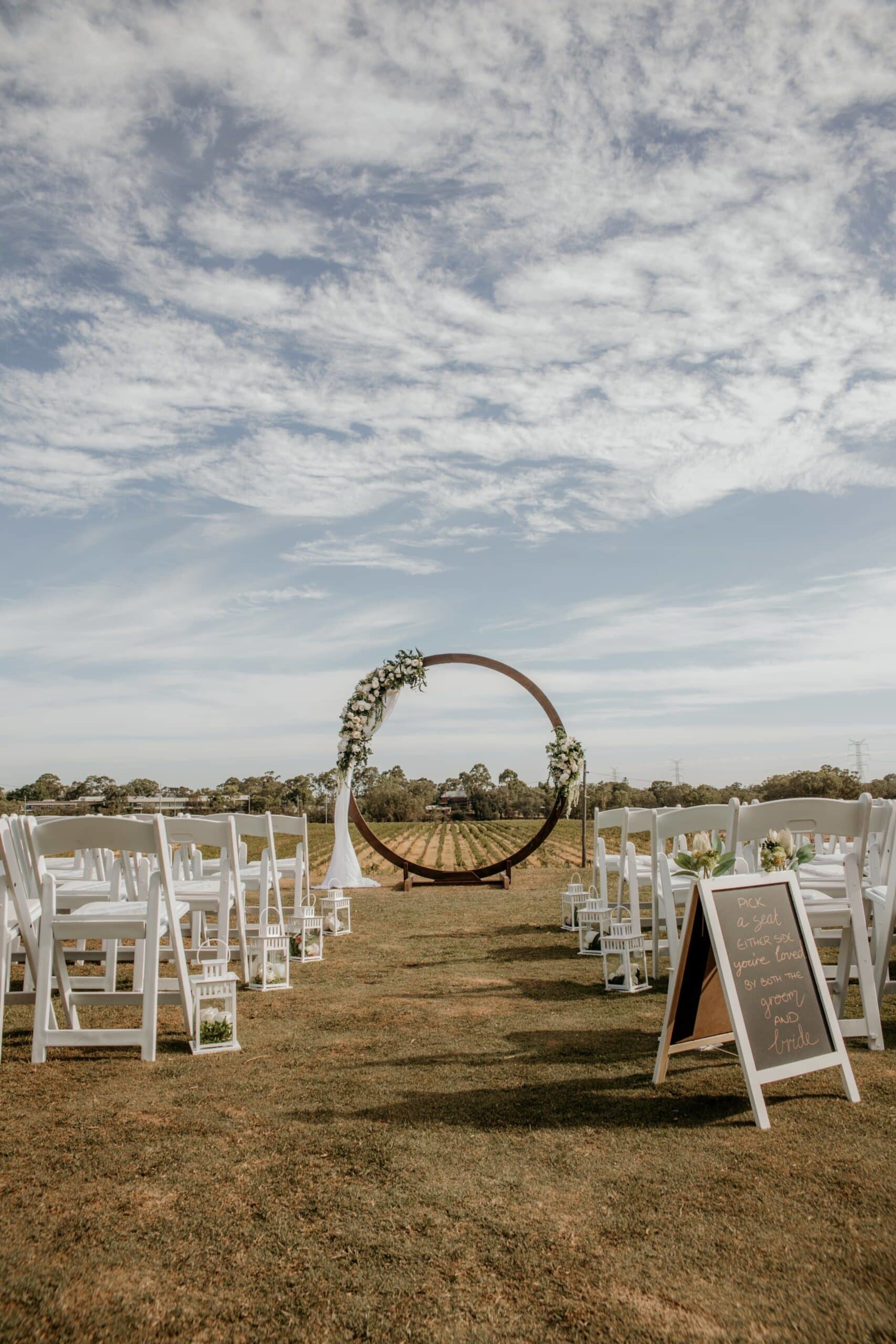 Outdoor vineyard wedding ceremony setup with white chairs and a circular floral arch under a cloudy sky.