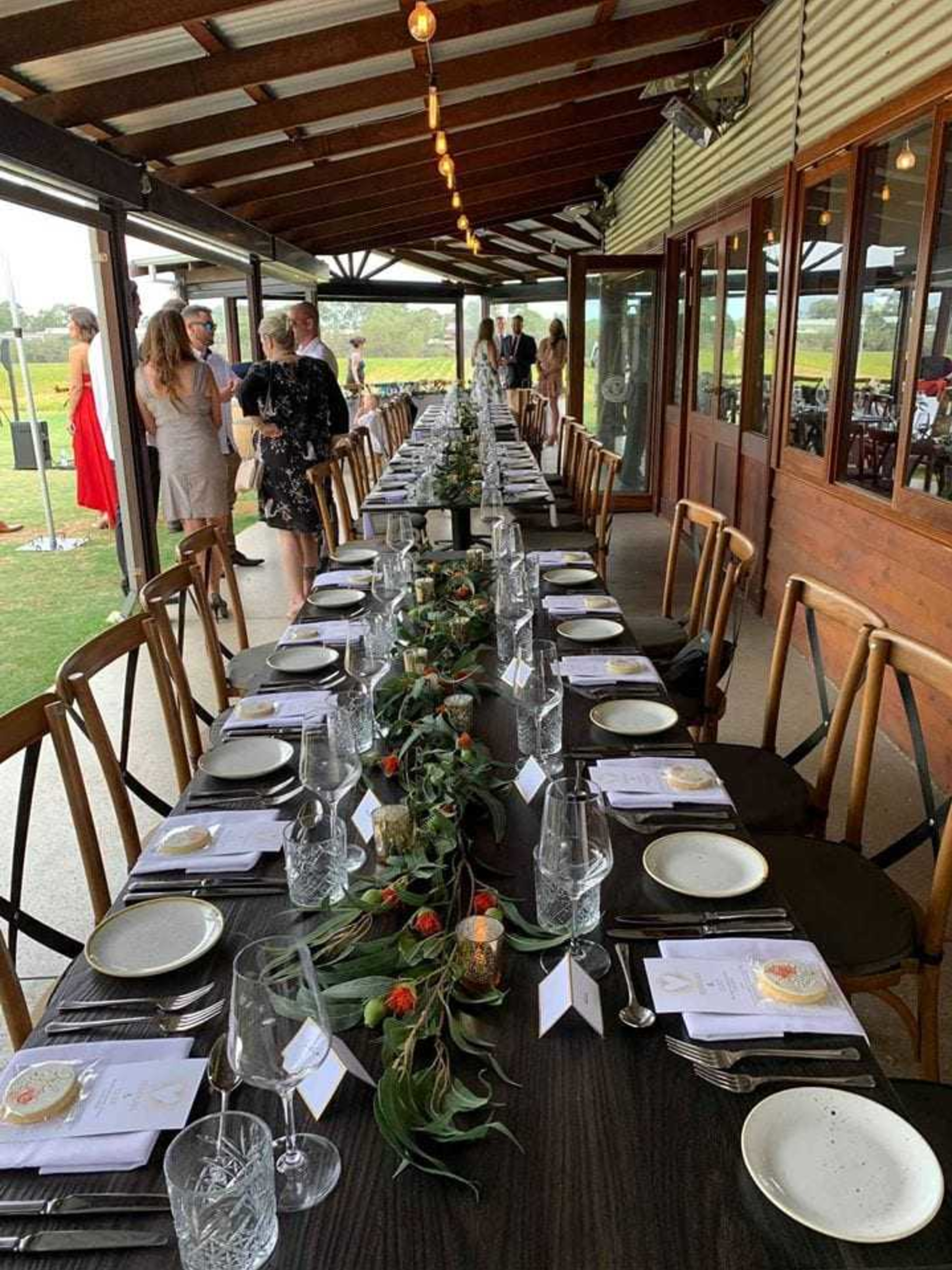 Rustic wedding reception with a long banquet table set with greenery garland under a covered outdoor pavilion.