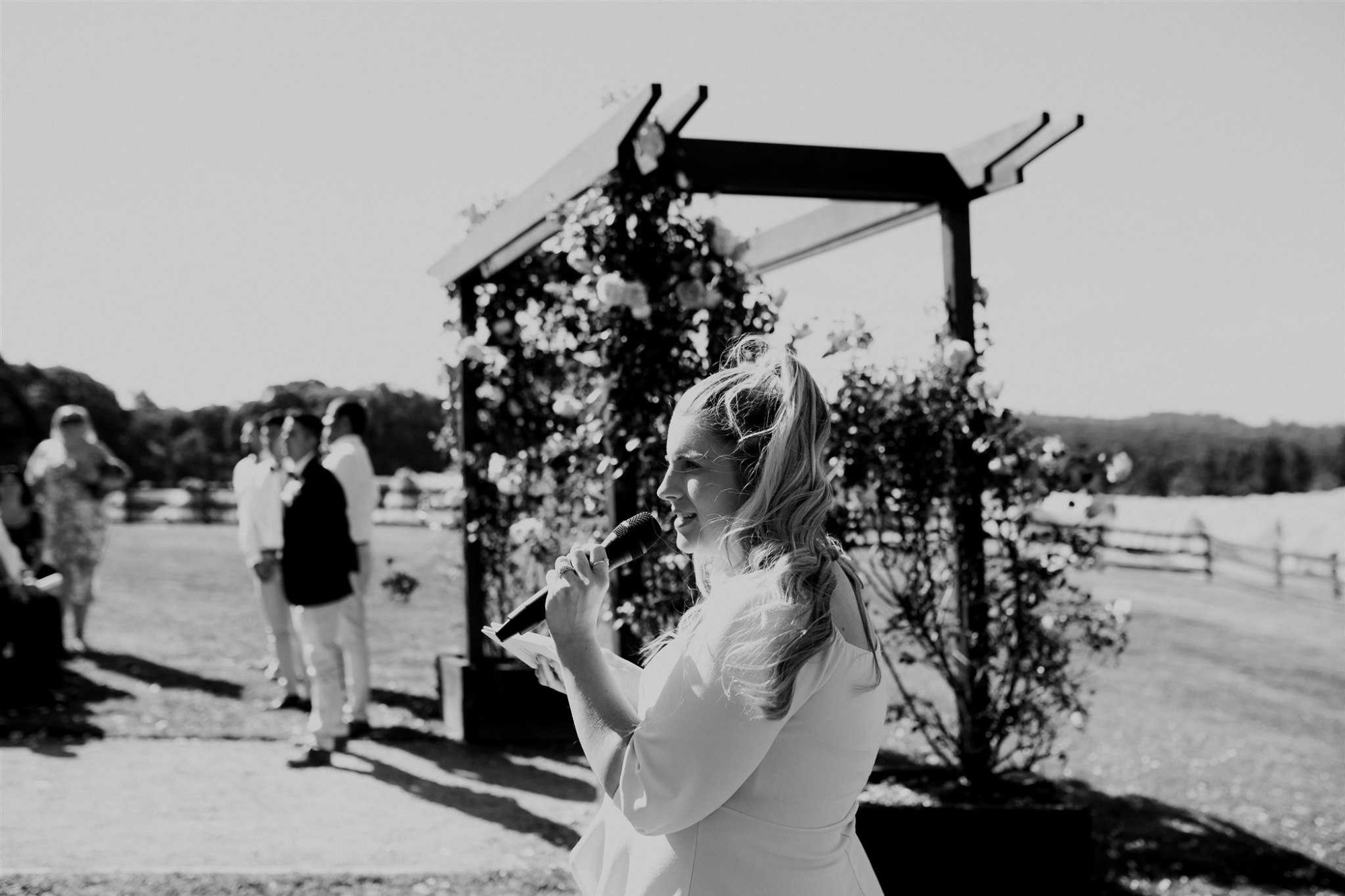 Wedding celebrant speaking into a microphone under a floral arch at an outdoor ceremony.