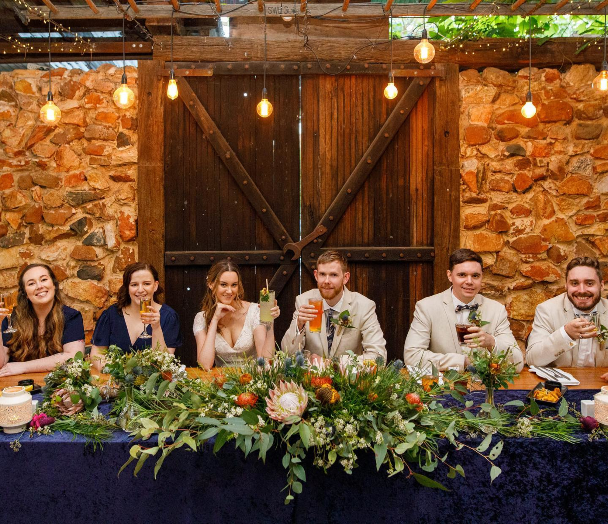 Wedding party seated at a rustic head table with lush greenery, drinks, and warm string lights against a stone wall backdrop.