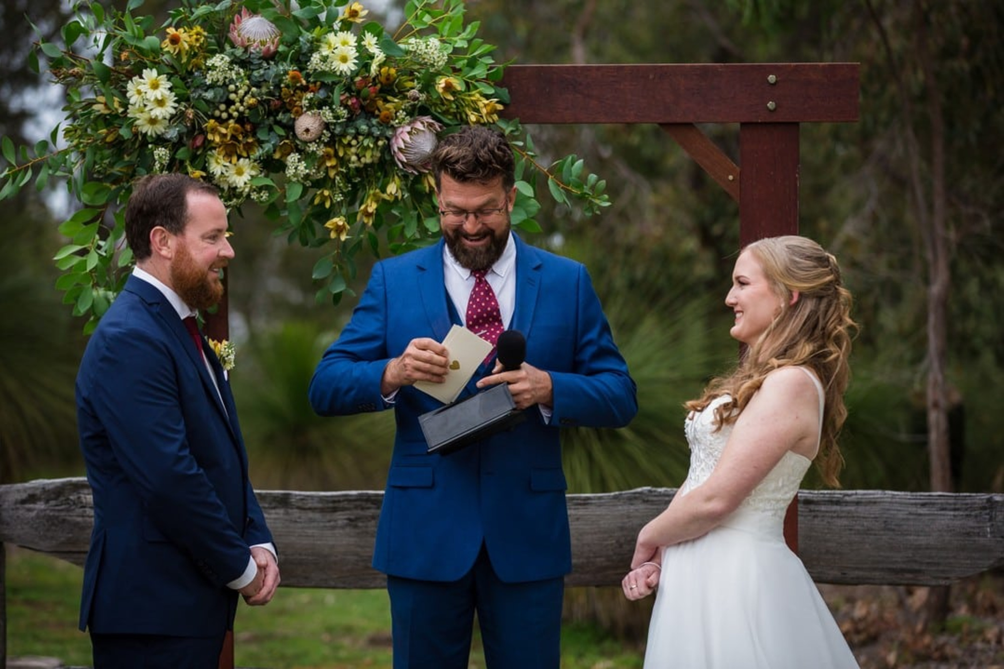 An officiant leads an outdoor rustic wedding ceremony between a smiling bride and groom under a floral arch.