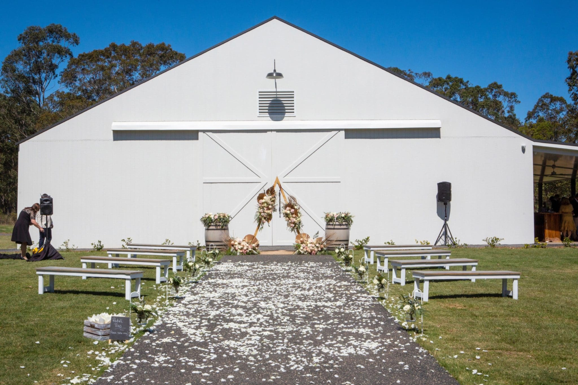 Outdoor wedding ceremony setup with benches, petal-strewn aisle, and floral arch in front of a white barn.