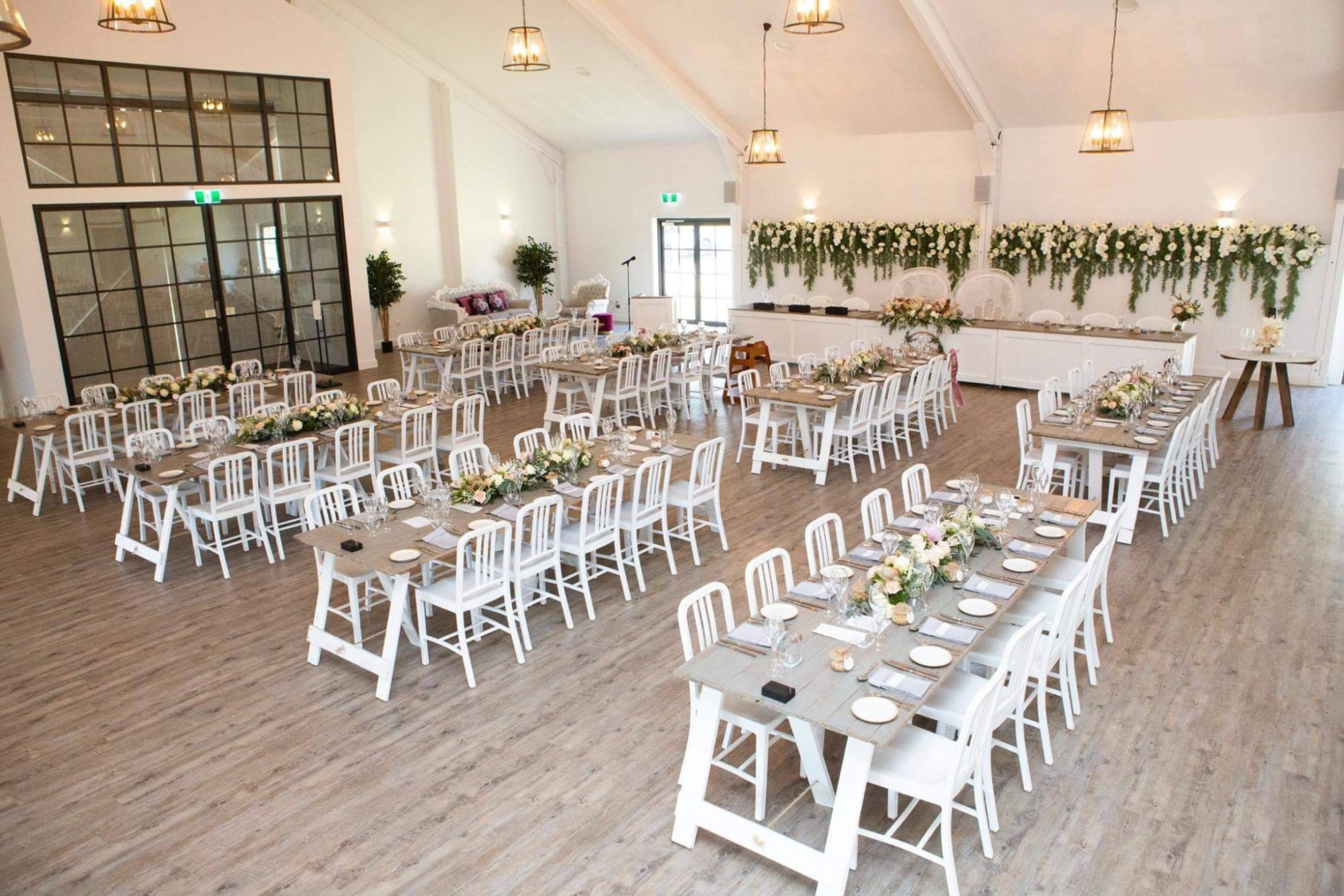 Bright indoor wedding reception venue with long white tables, chairs, and lush floral greenery decor.