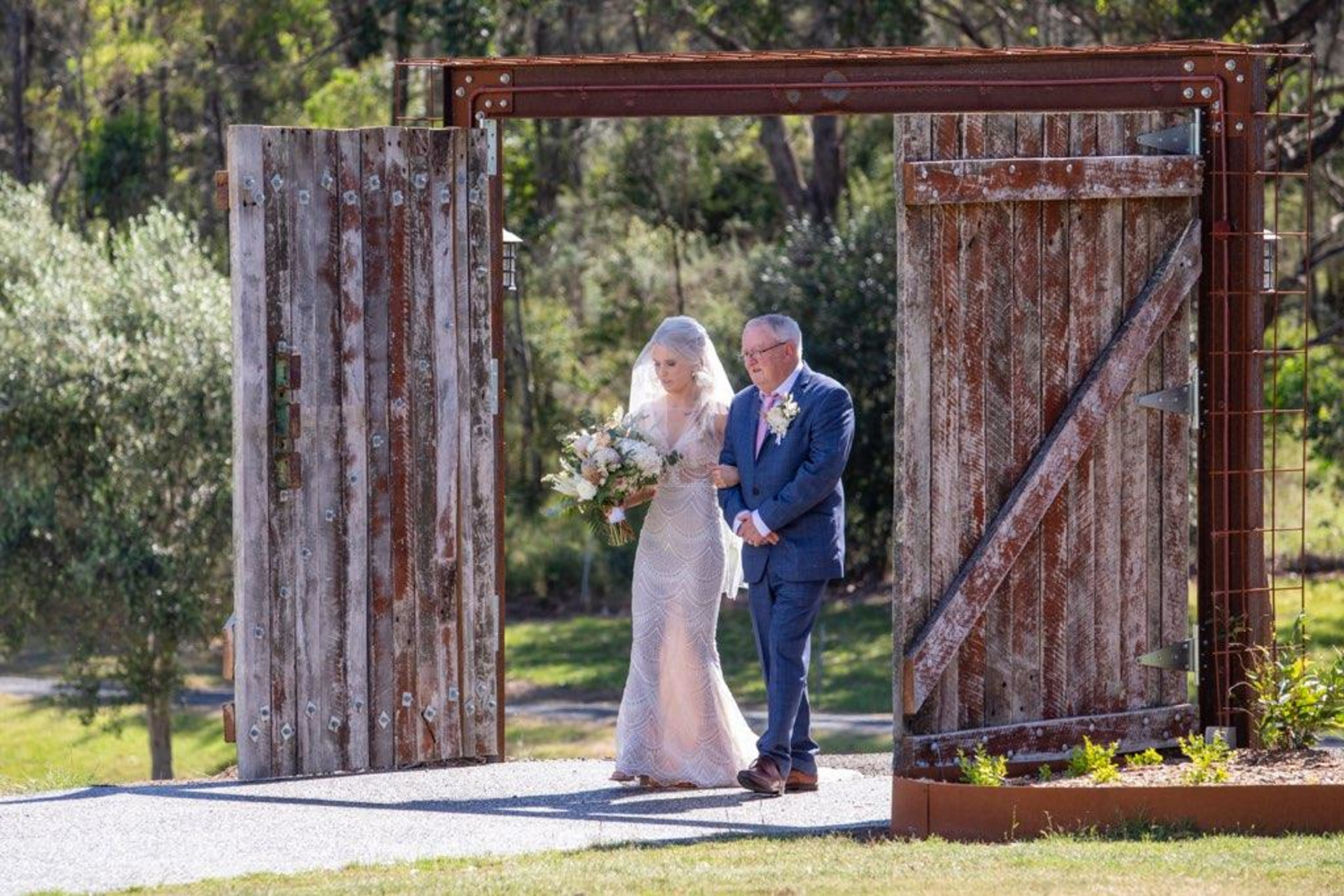 Bride with bouquet walks arm-in-arm with escort through large rustic wooden doors at an outdoor wedding venue.