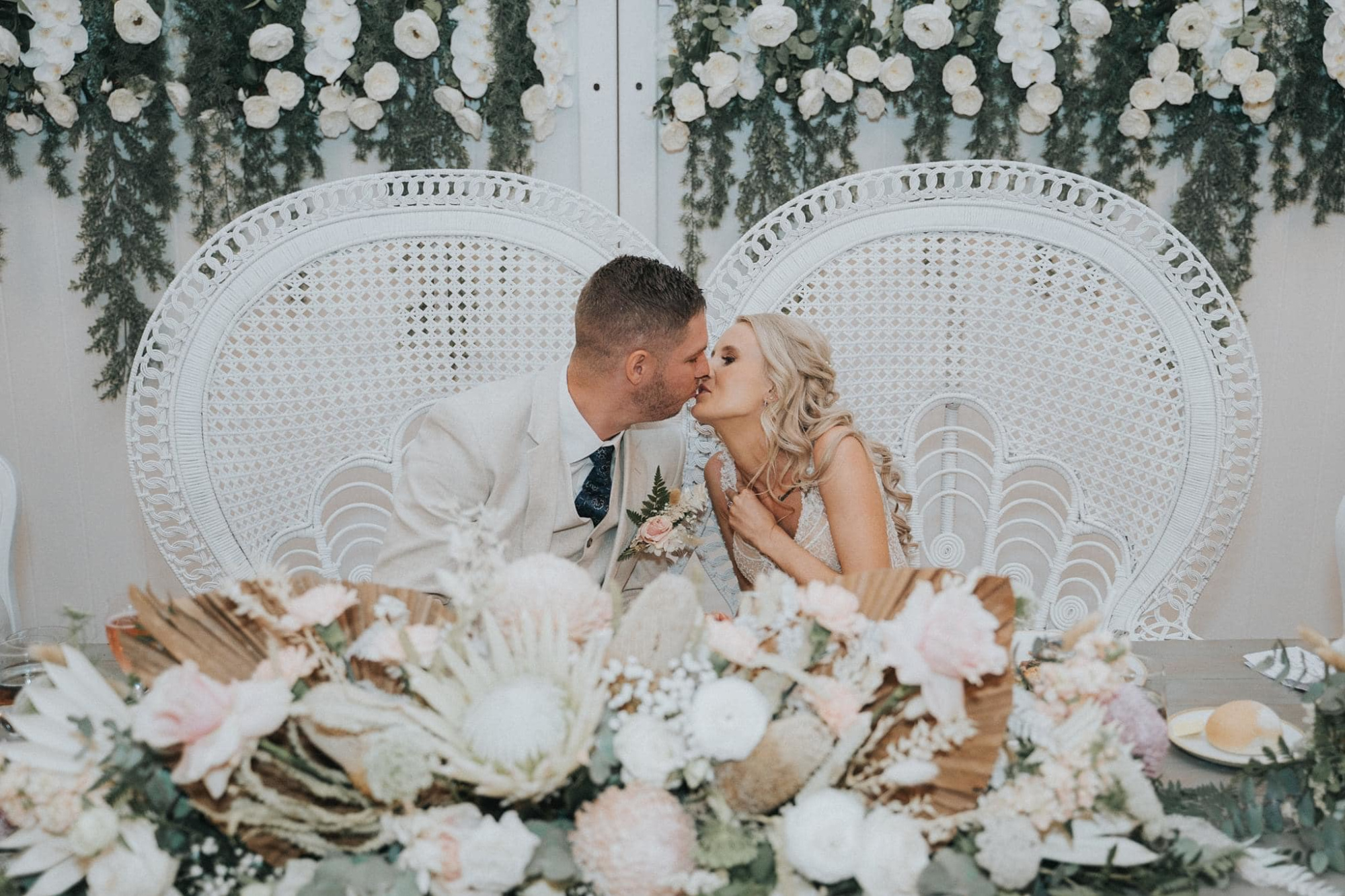 Bride and groom share a kiss at a boho sweetheart table with white wicker chairs and lush floral backdrop.