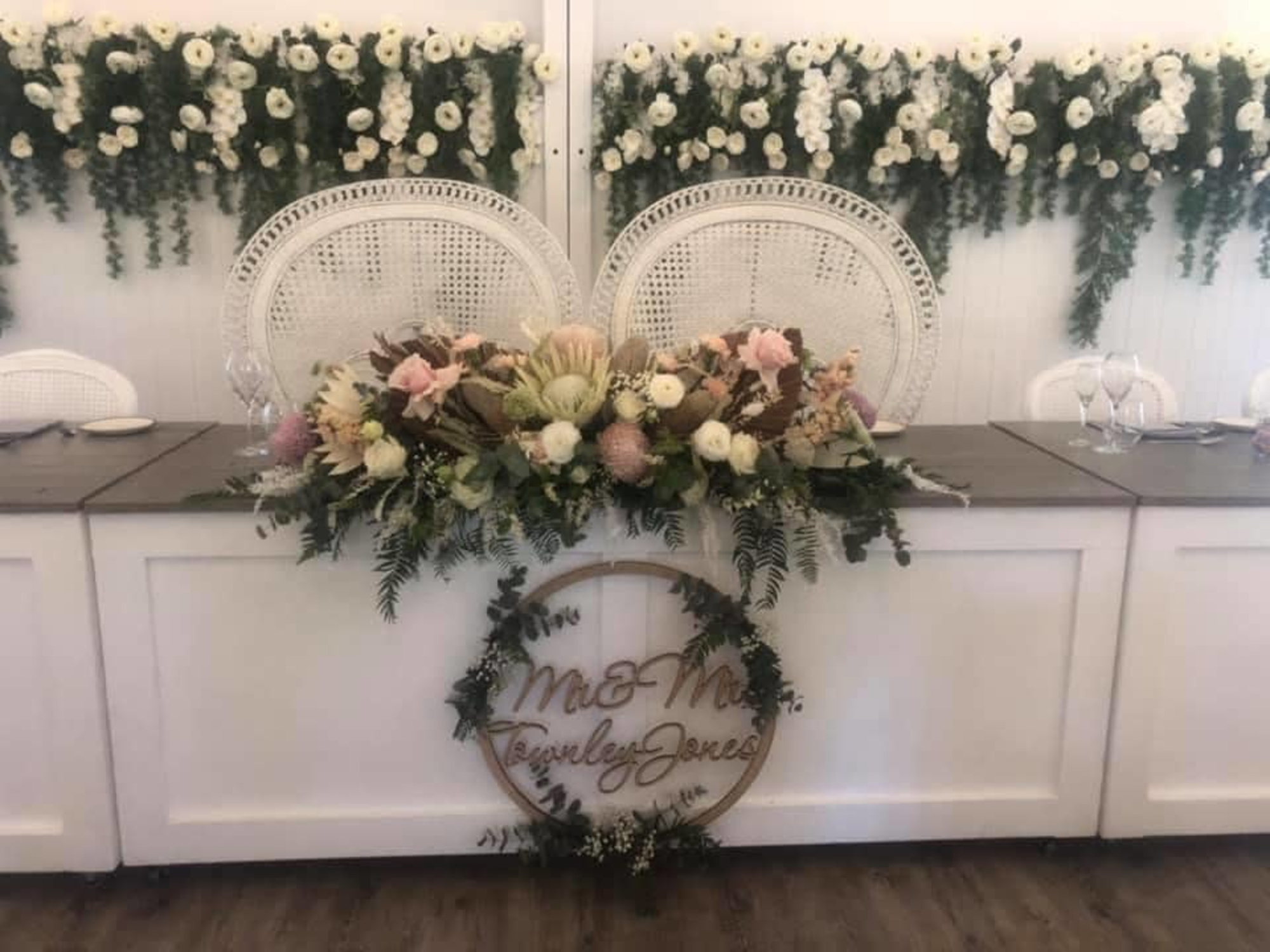 Boho sweetheart table with large floral centerpiece, rattan chairs, and a greenery flower backdrop at a wedding reception.