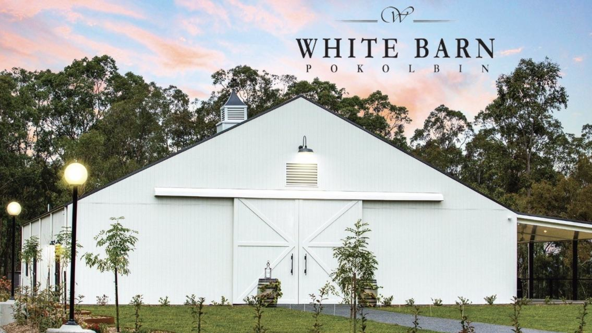 White barn wedding venue surrounded by trees at sunset with landscaped garden path.