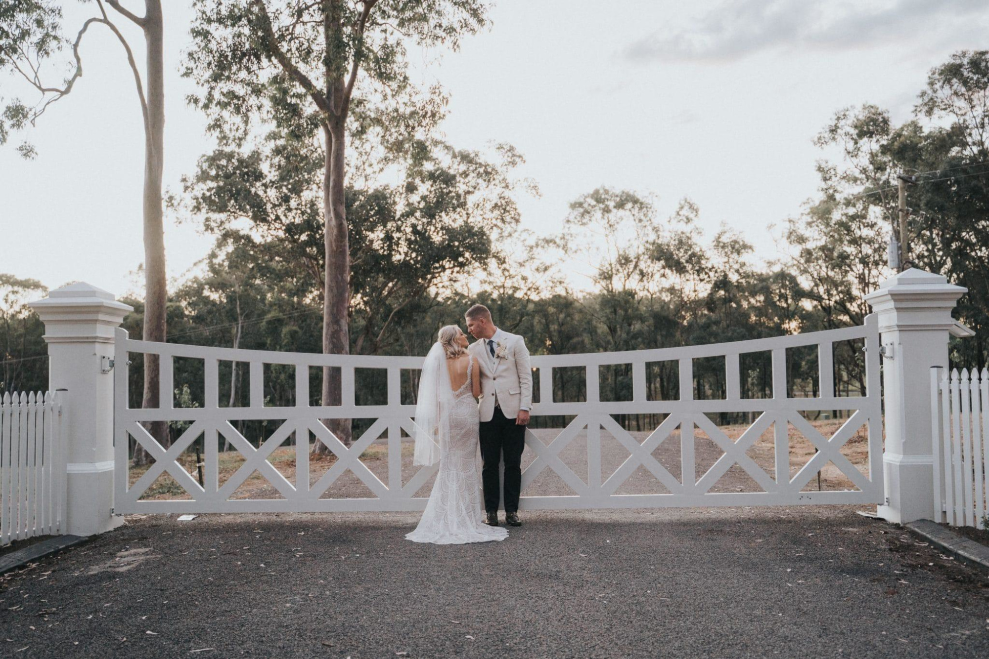 Bride and groom embrace in front of a white country gate at sunset.