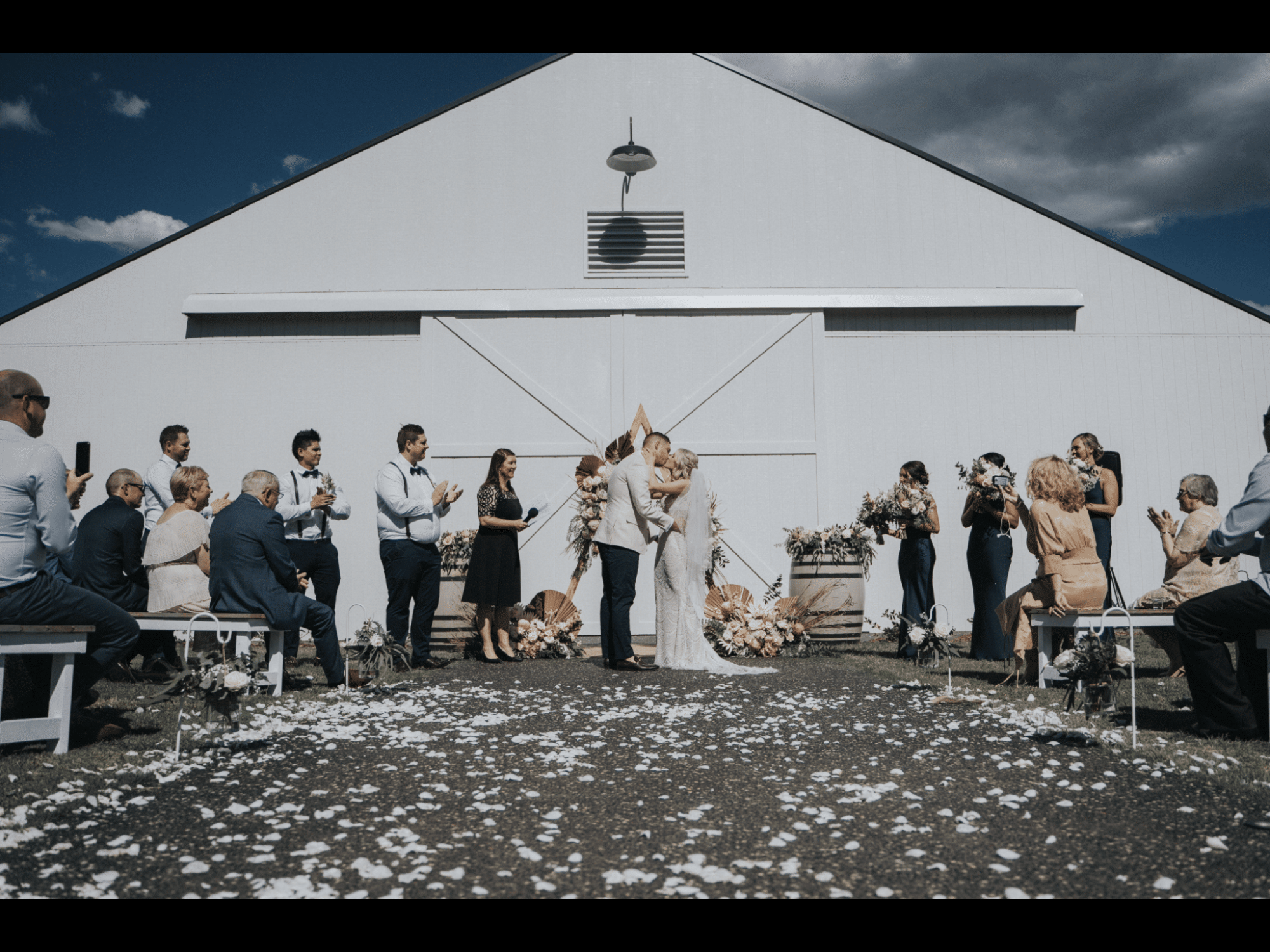 Outdoor wedding ceremony in front of a white barn as the couple kisses surrounded by guests and floral decor.
