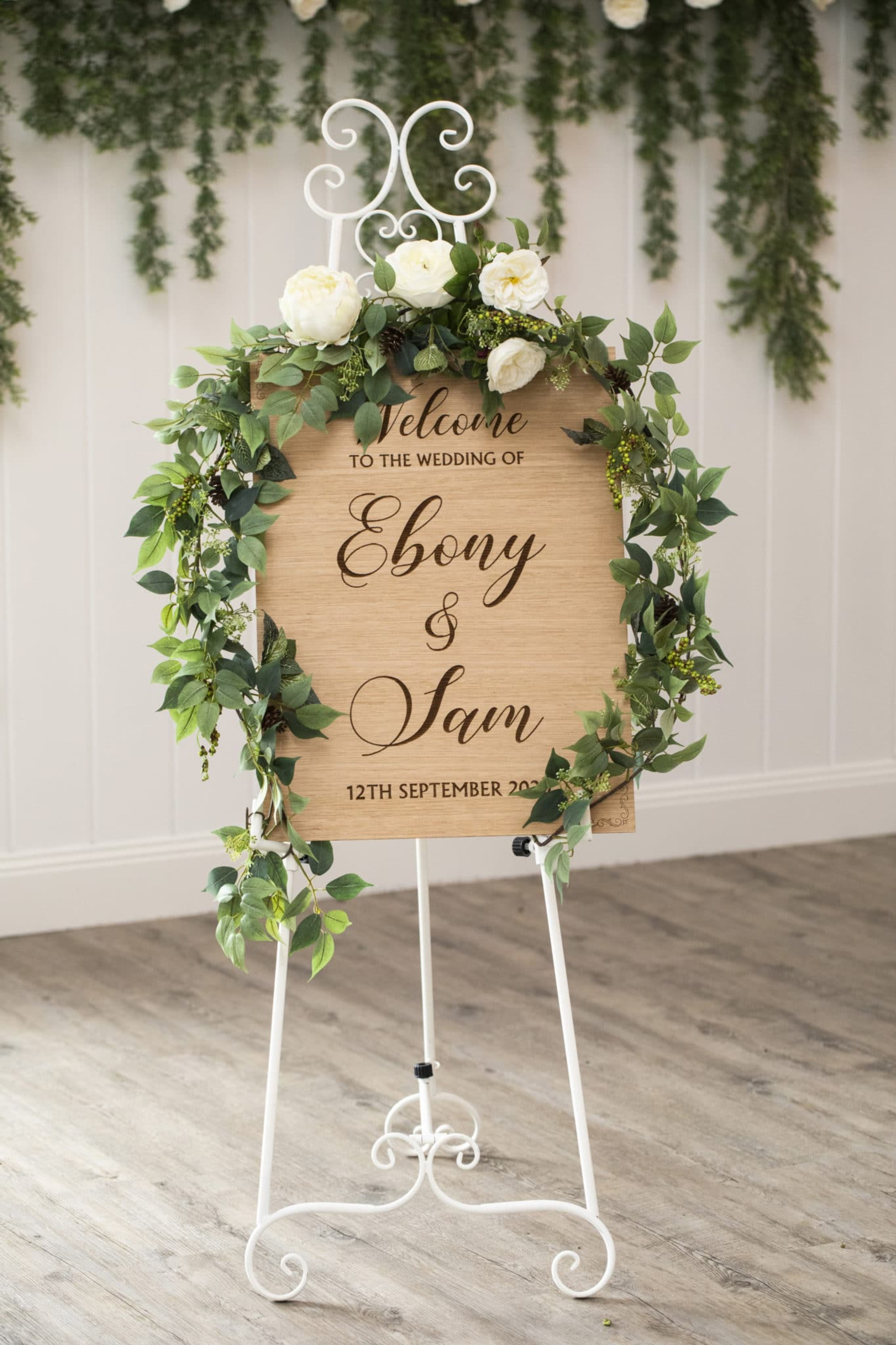 Rustic wooden wedding welcome sign with greenery and white flowers on a white metal easel.