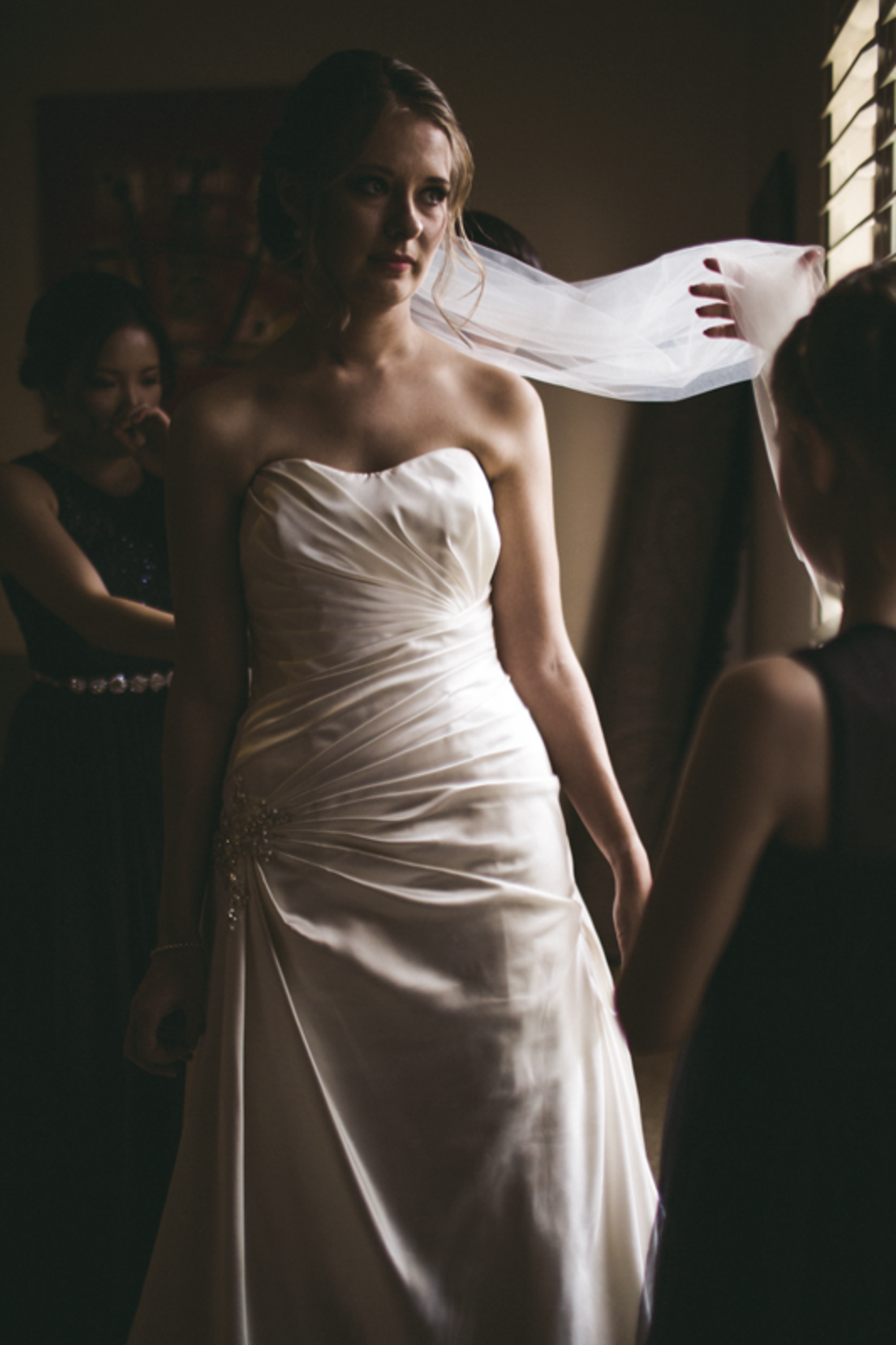 Bride in a strapless gown stands by a window as her bridesmaids adjust her veil in soft, moody light.