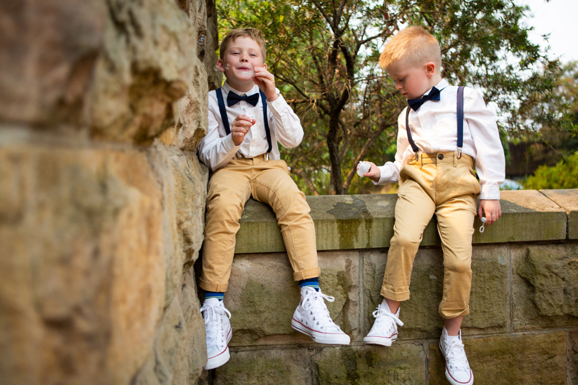 Two young boys in suspenders and bow ties sit on a stone wall blowing bubbles at an outdoor wedding.