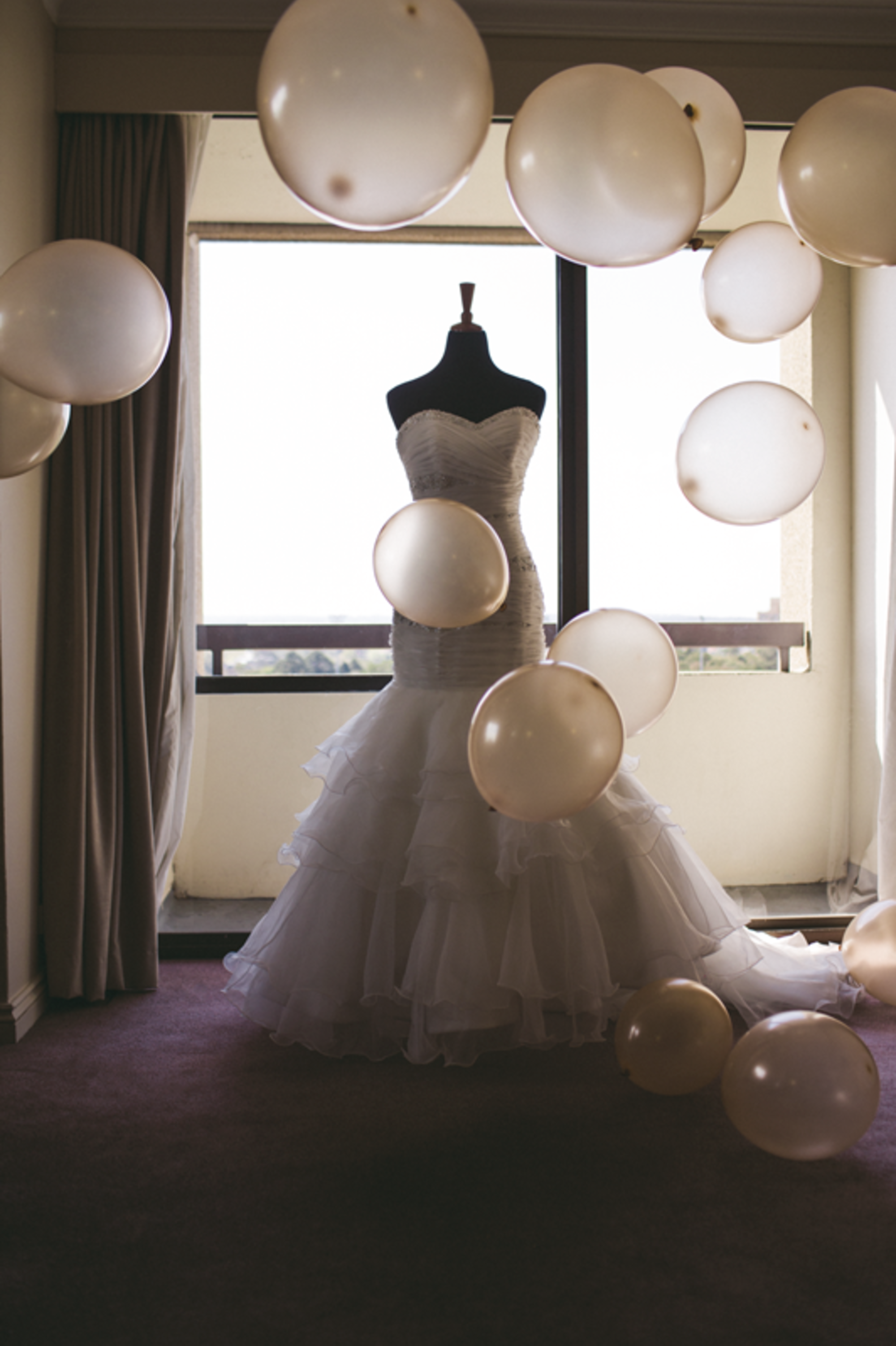 Strapless ruffled wedding dress on a mannequin surrounded by floating balloons in front of a bright window.