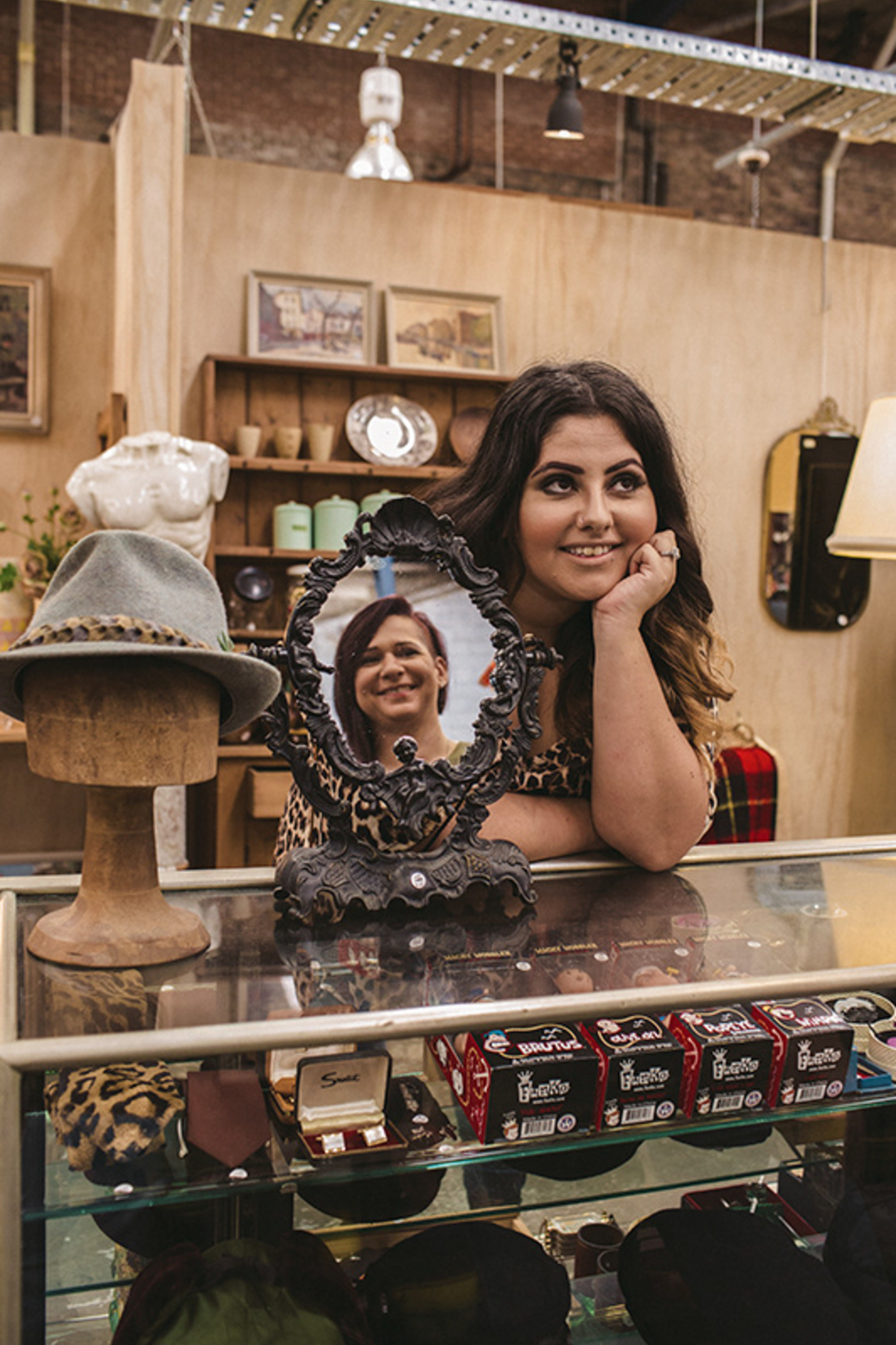 Woman in a vintage shop leaning on a glass counter with an ornate mirror and retro decor around her.