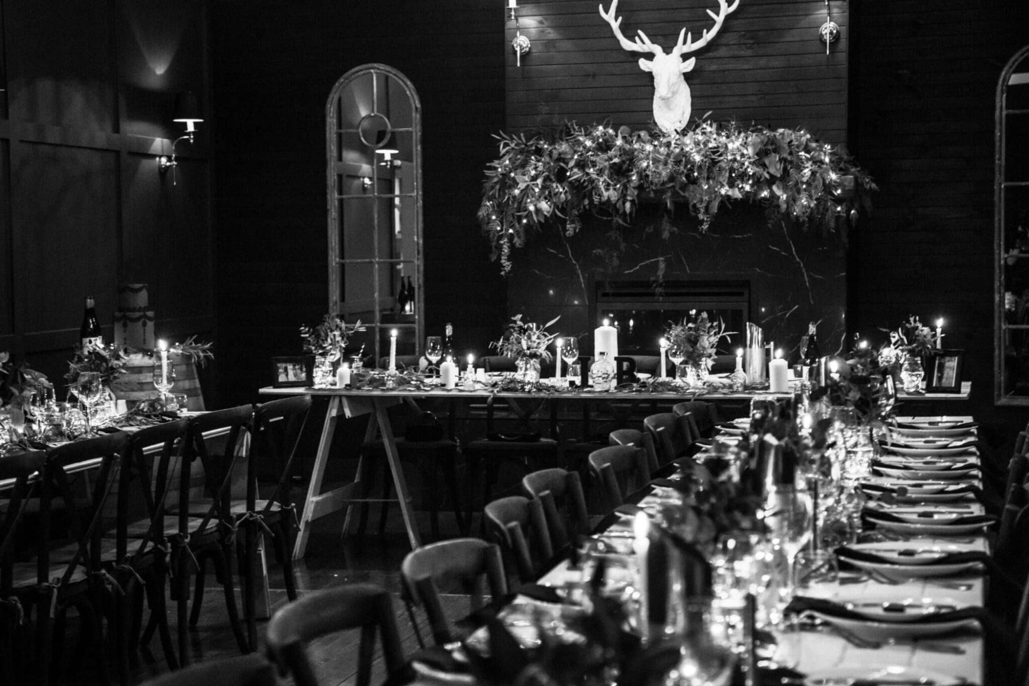 Candlelit wedding reception with long rustic tables, greenery garlands, and a stag mount above a dark fireplace.