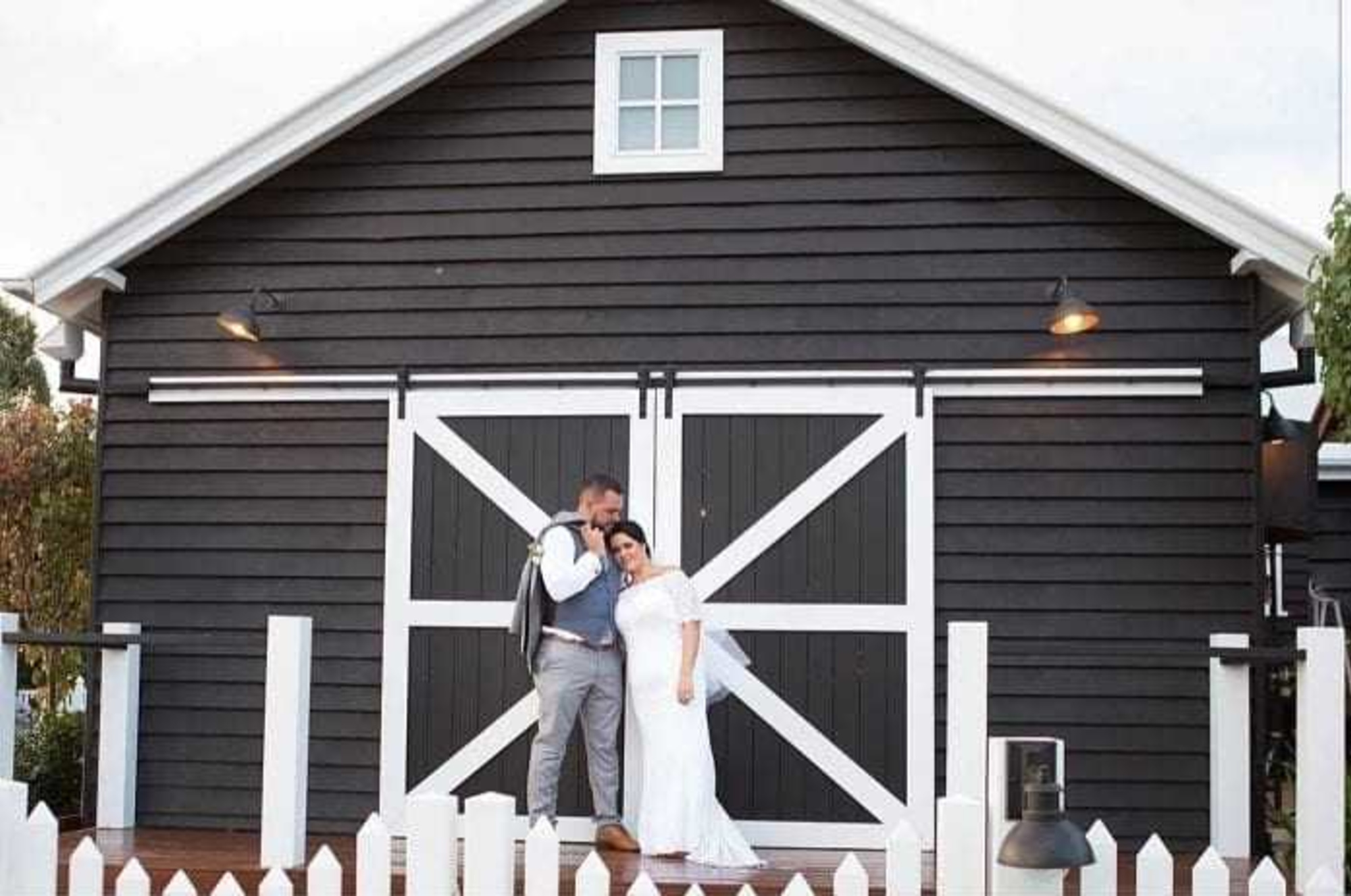 Wedding couple embraces in front of a modern black and white barn with a white picket fence.