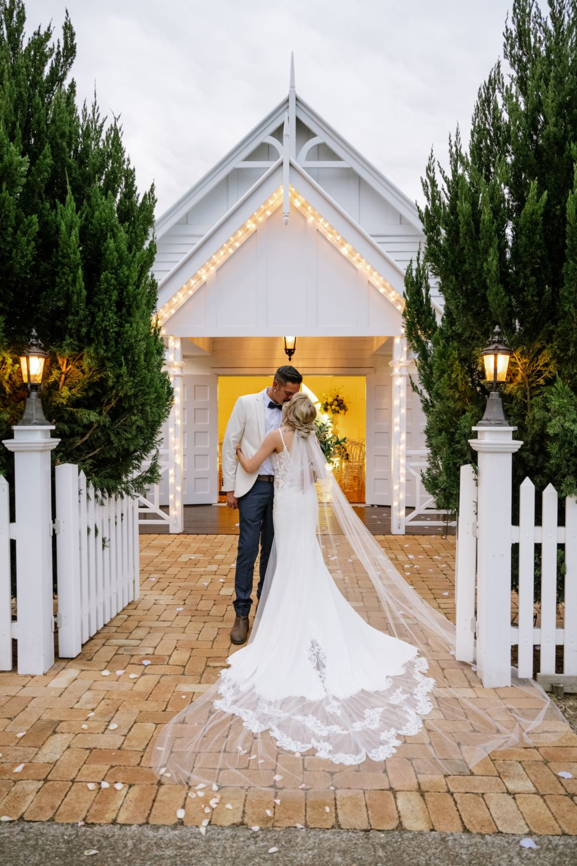 Bride and groom share a kiss in front of a lit white chapel framed by greenery and a picket fence.