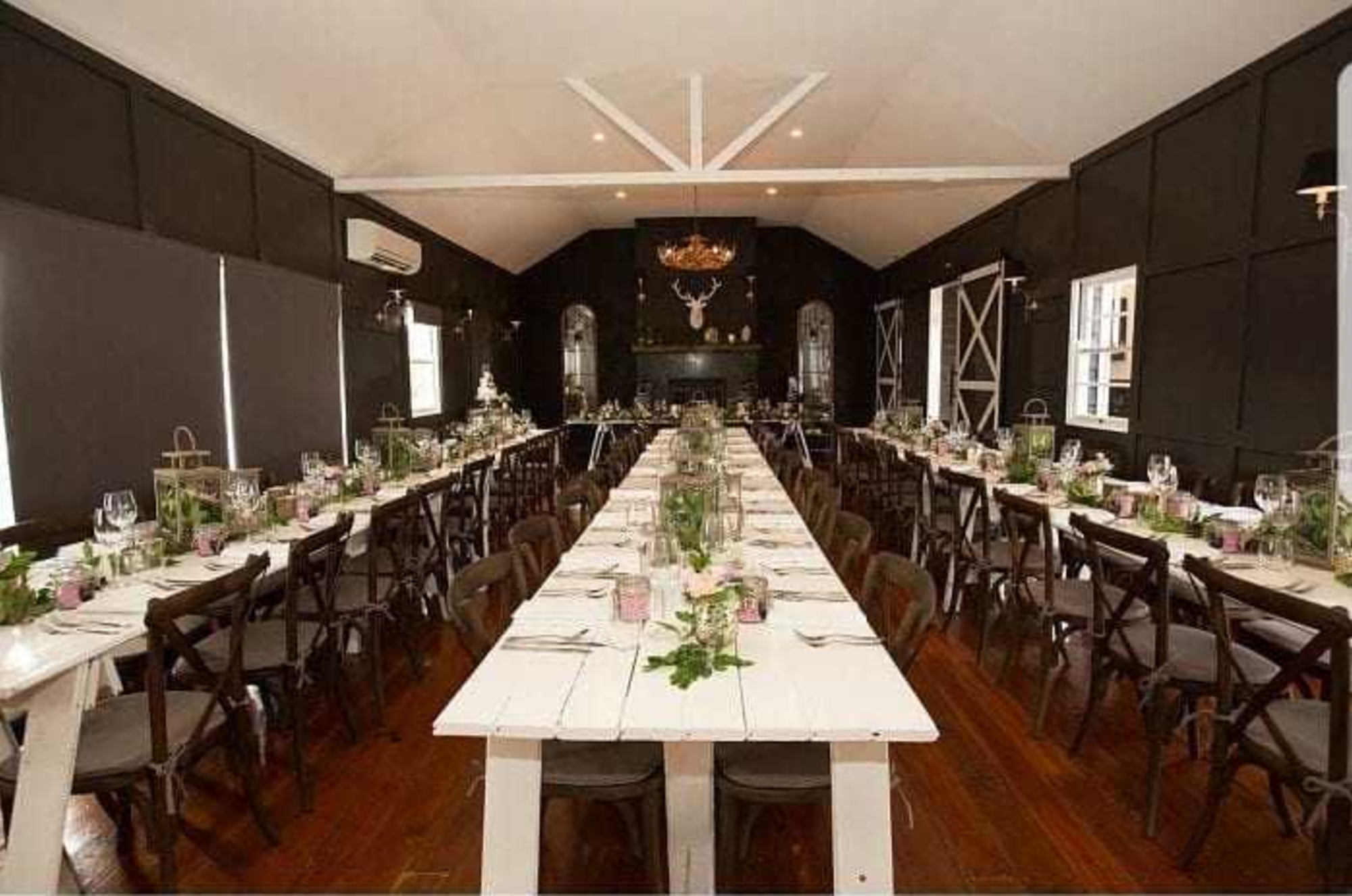 Rustic indoor wedding reception with long white banquet tables, dark chairs, and greenery centerpieces.