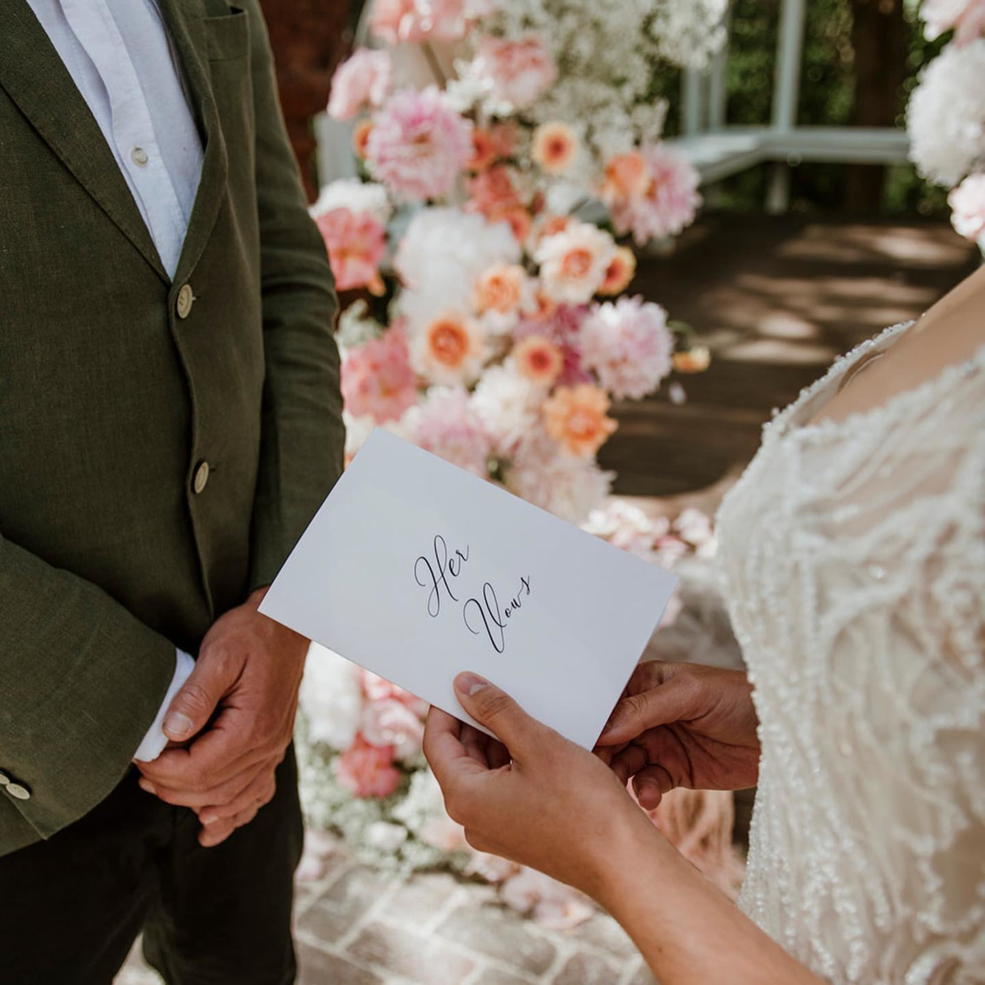 Bride holding a vow booklet while facing the groom in front of a romantic floral wedding backdrop.