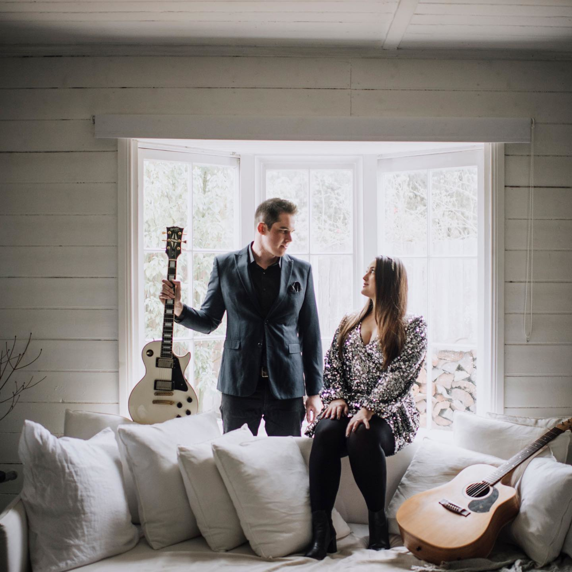 Stylish musician couple with guitars posing on a white sofa in front of bright bay windows.