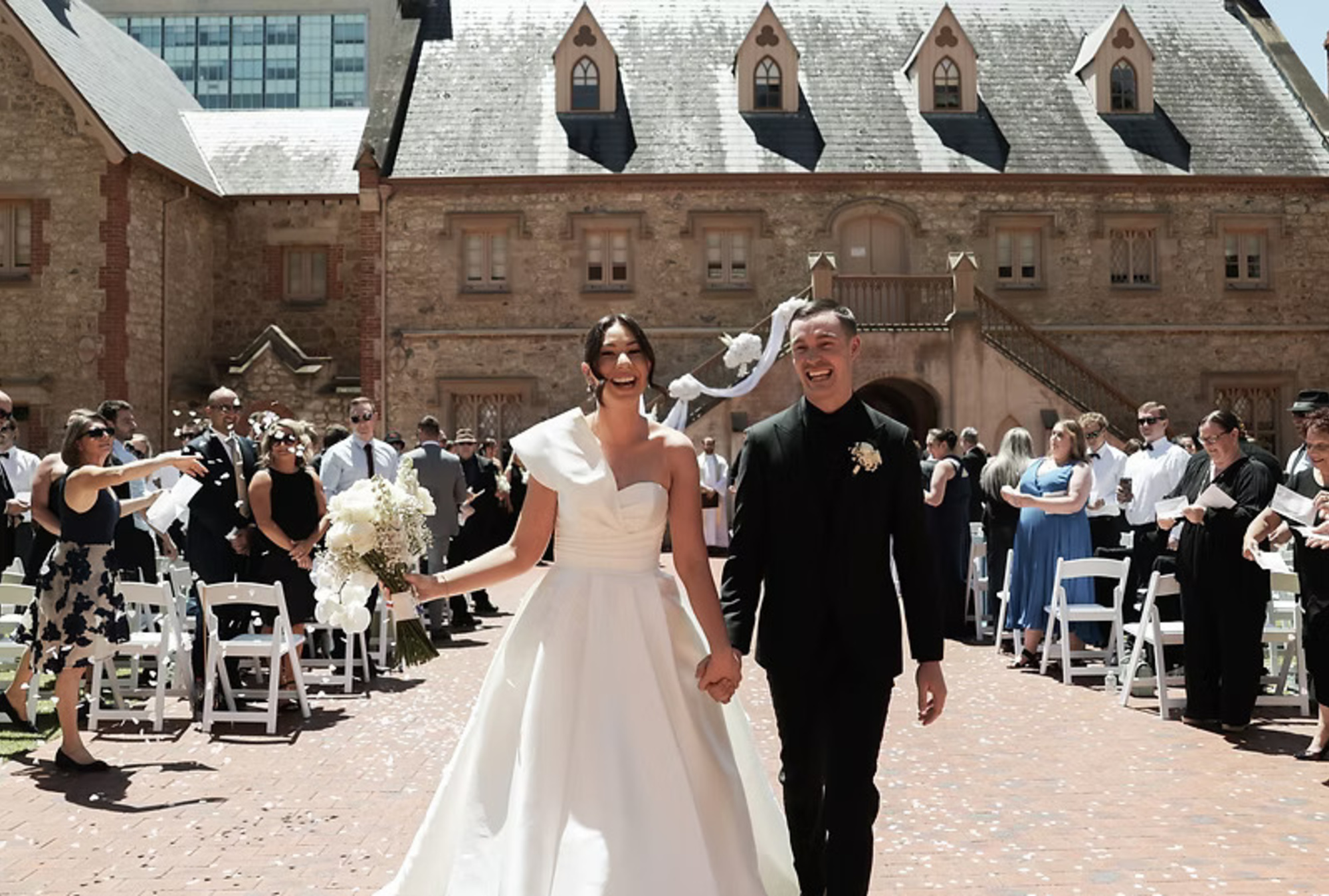 Joyful bride and groom walk back up the aisle outdoors surrounded by guests in a historic courtyard venue.