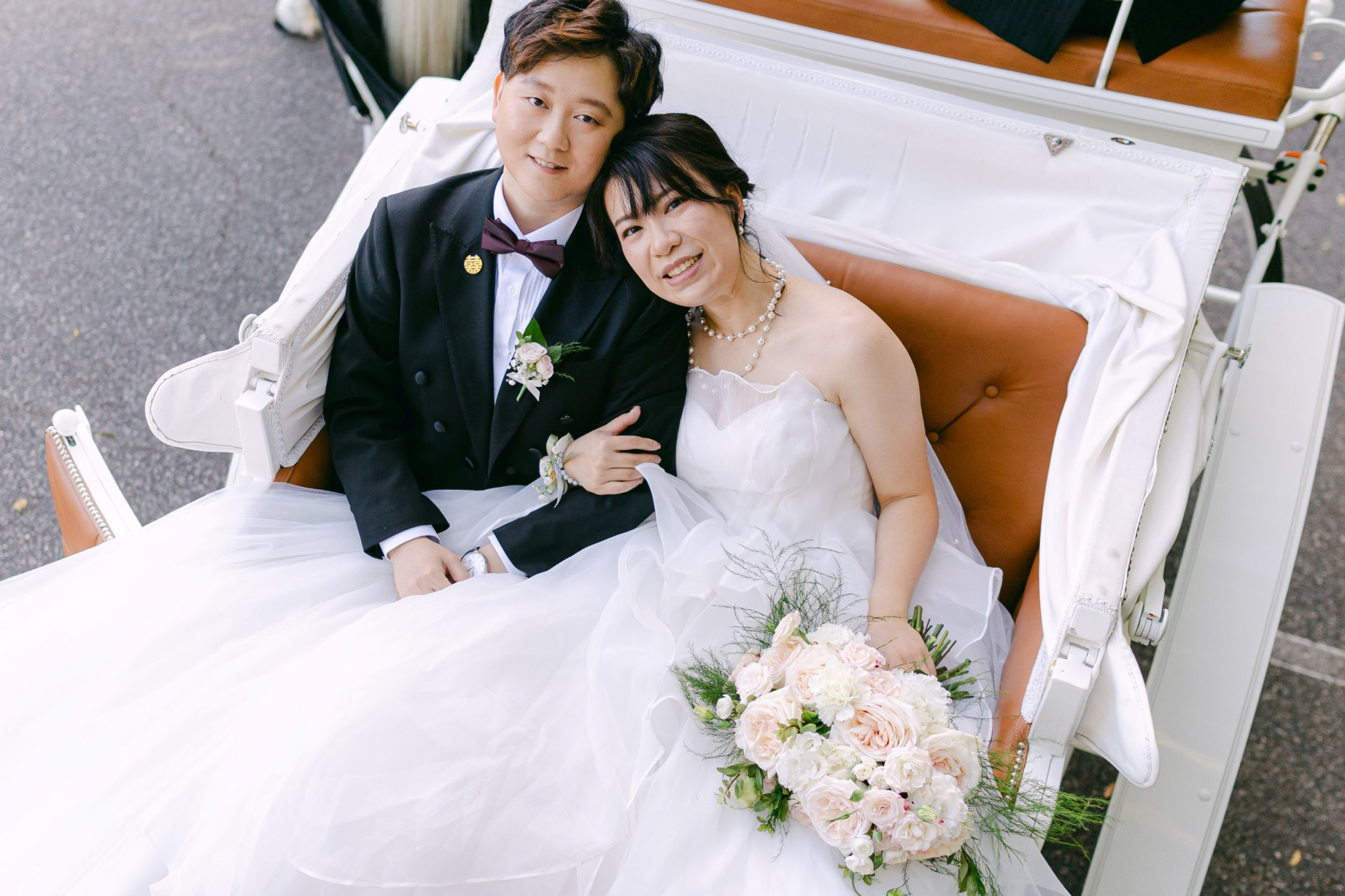 Smiling wedding couple in formal attire sitting together in a white carriage holding a pastel rose bouquet.
