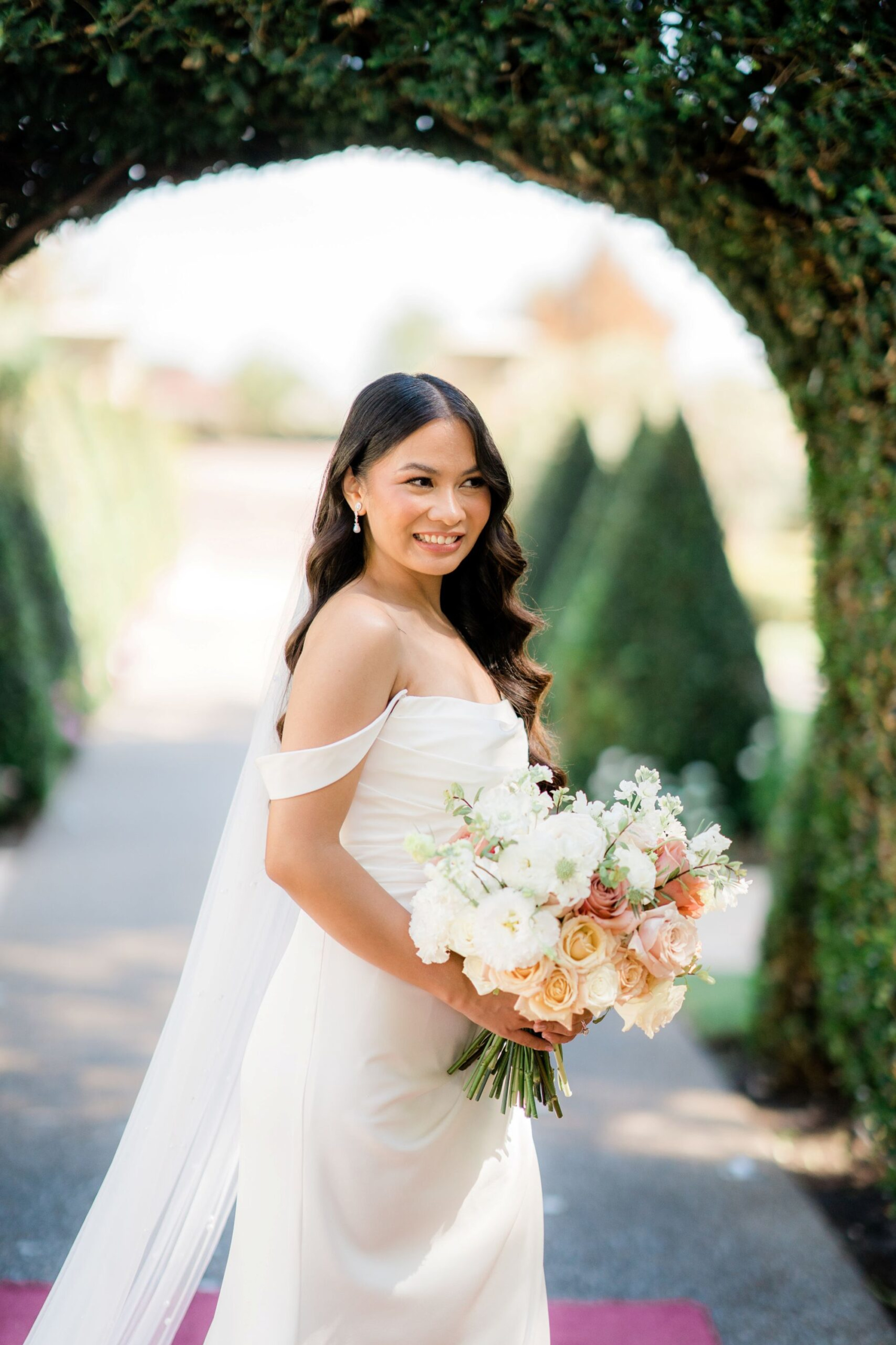 Bride in an off-the-shoulder gown holding a pastel bouquet under a lush green garden archway.