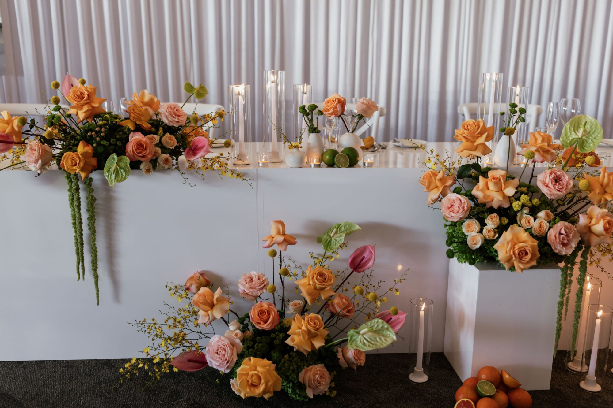 Modern wedding reception table with peach and pink floral arrangements, candles, and citrus accents against a white backdrop.