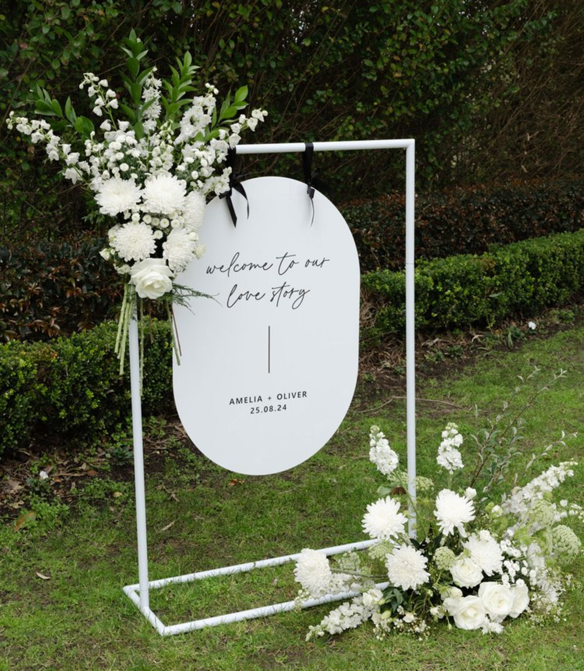 Minimalist white wedding welcome sign with black script and white floral arrangements on a metal stand in a garden.
