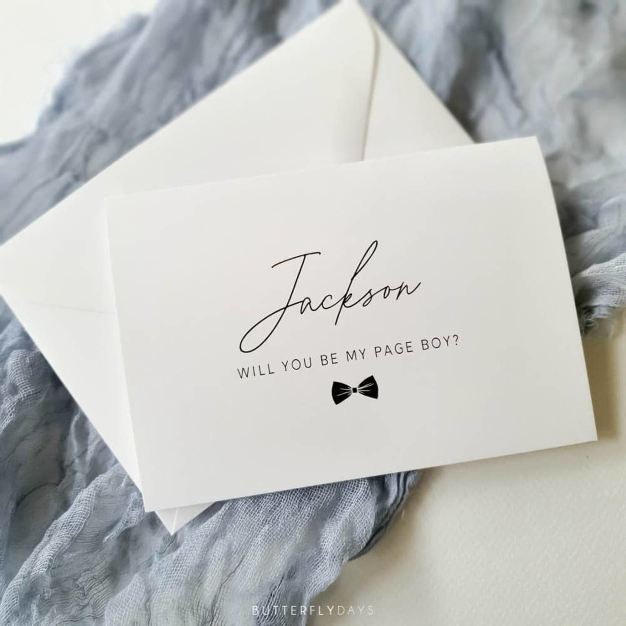 Minimalist page boy proposal card with elegant script text and bow tie icon on a white envelope over soft grey fabric.