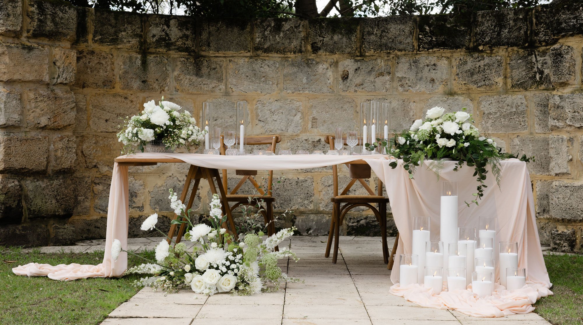 Outdoor wedding sweetheart table with white florals, candles, and blush draping against a stone wall backdrop.