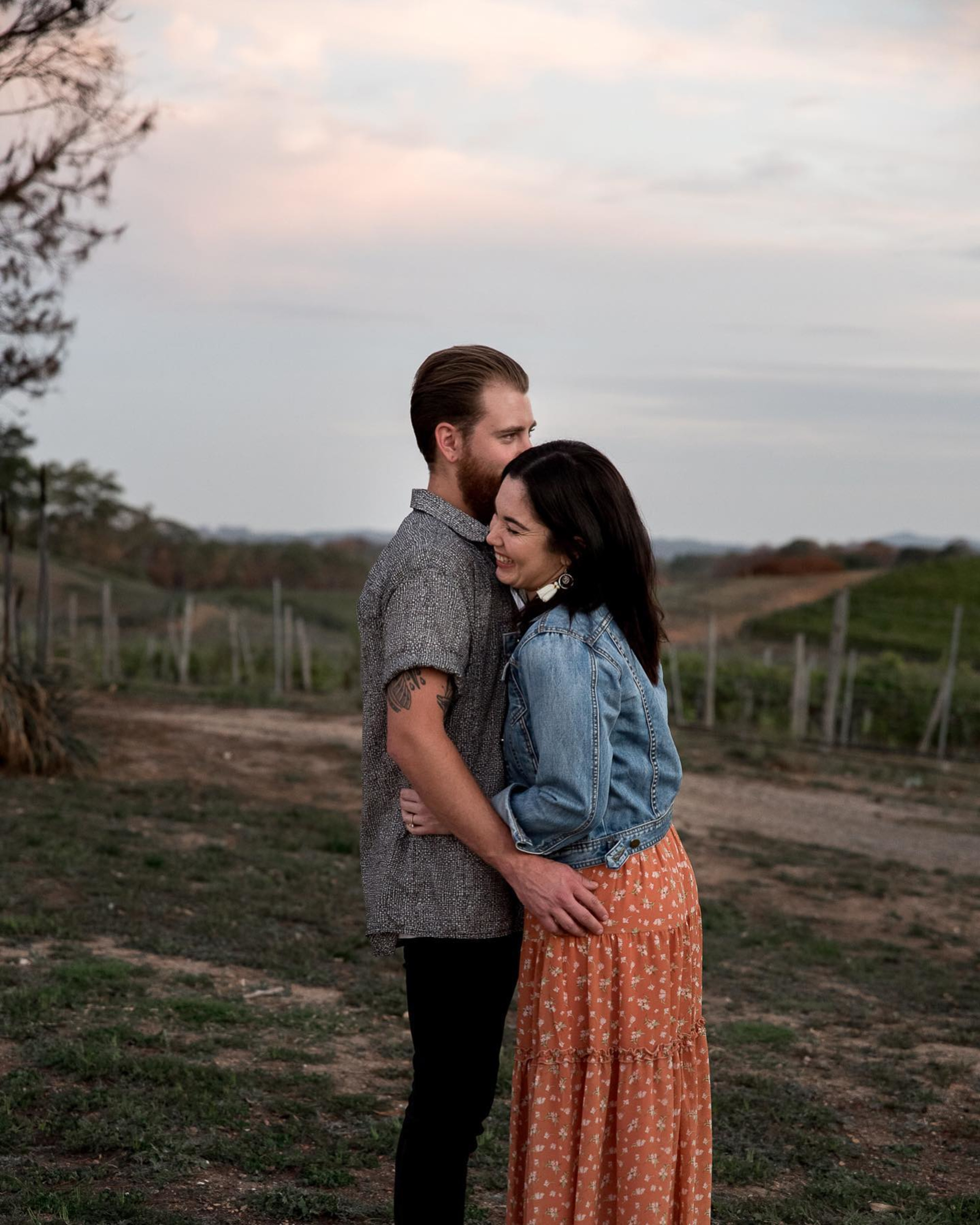 A couple embraces and smiles together outdoors at sunset in a rustic field.