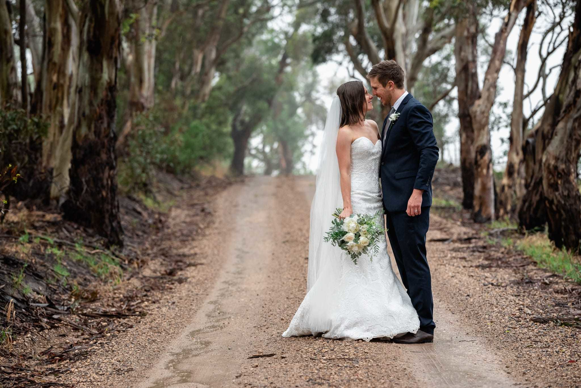 Bride and groom embrace on a rustic tree-lined dirt road holding a white and green bouquet.