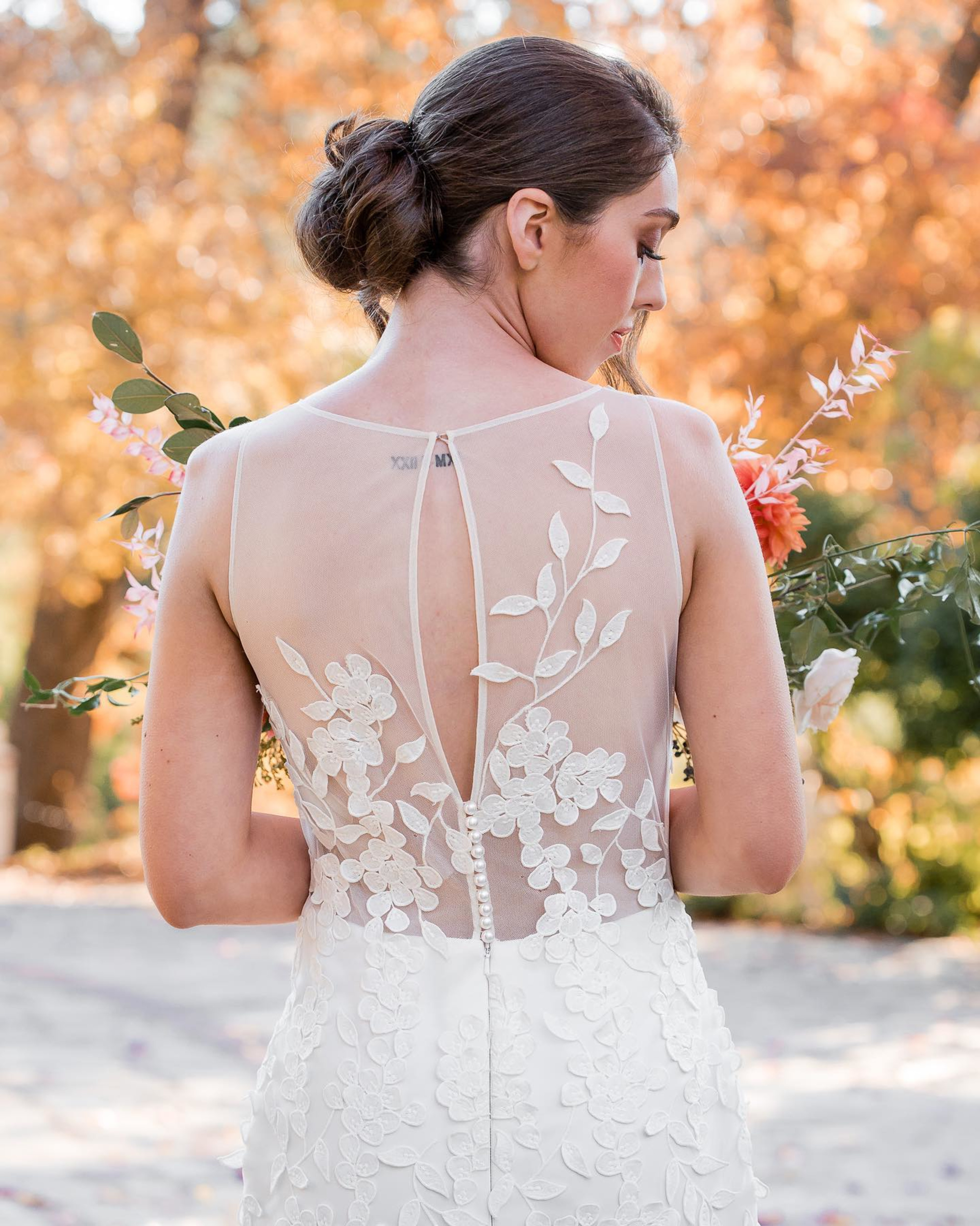 Bride in a lace-back wedding dress holding flowers in an outdoor setting with autumn foliage.