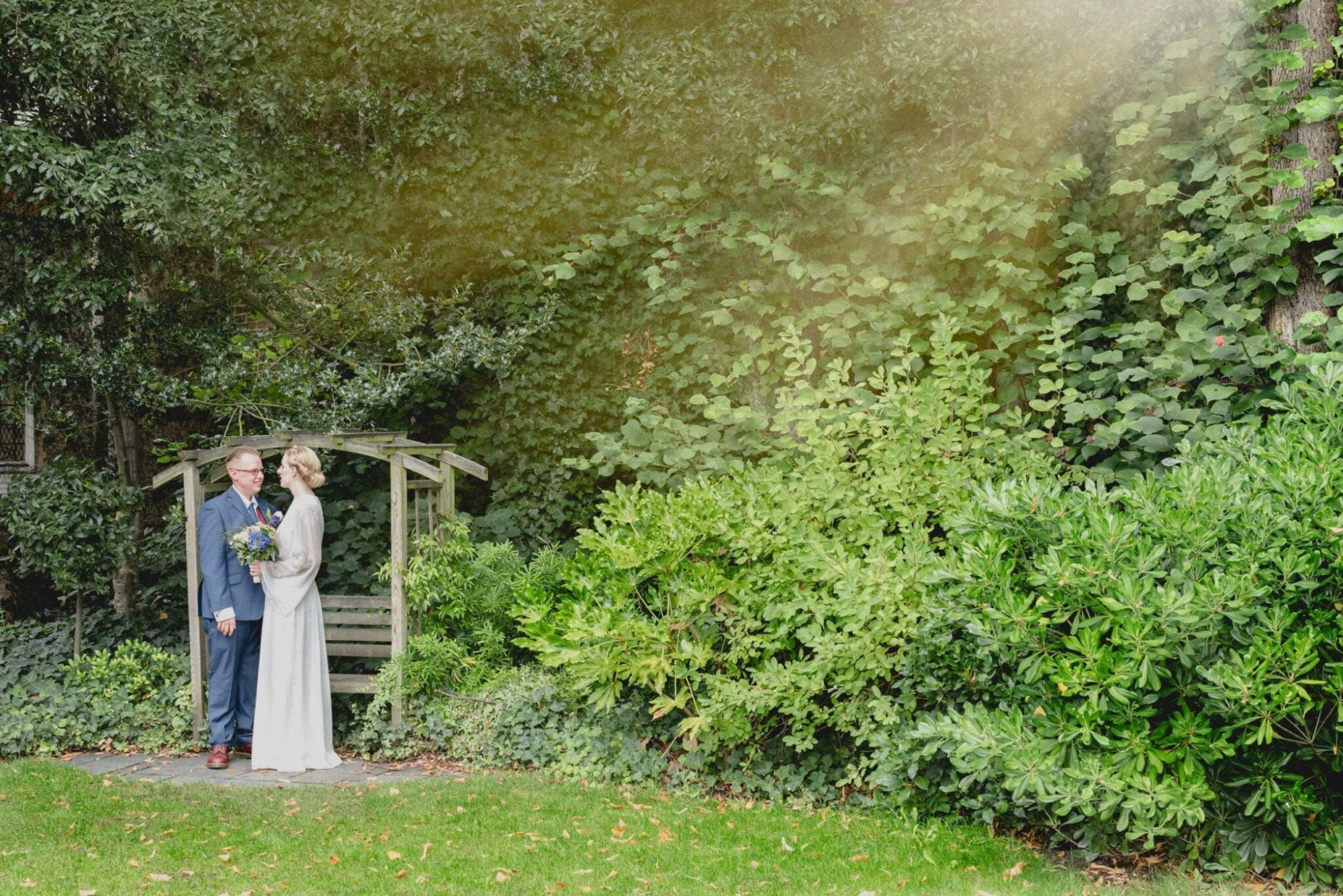 A wedding couple stands together under a wooden garden arch surrounded by lush green foliage.