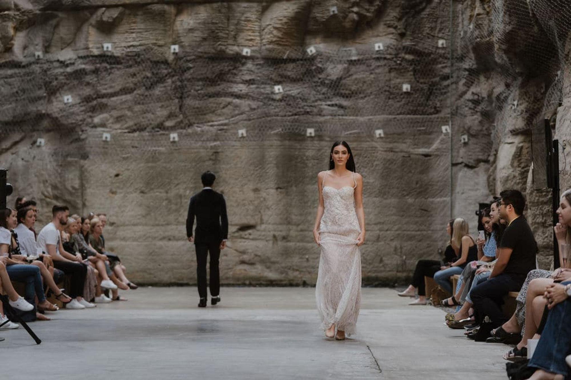 Bride walks down an industrial runway in a lace wedding gown surrounded by seated guests.