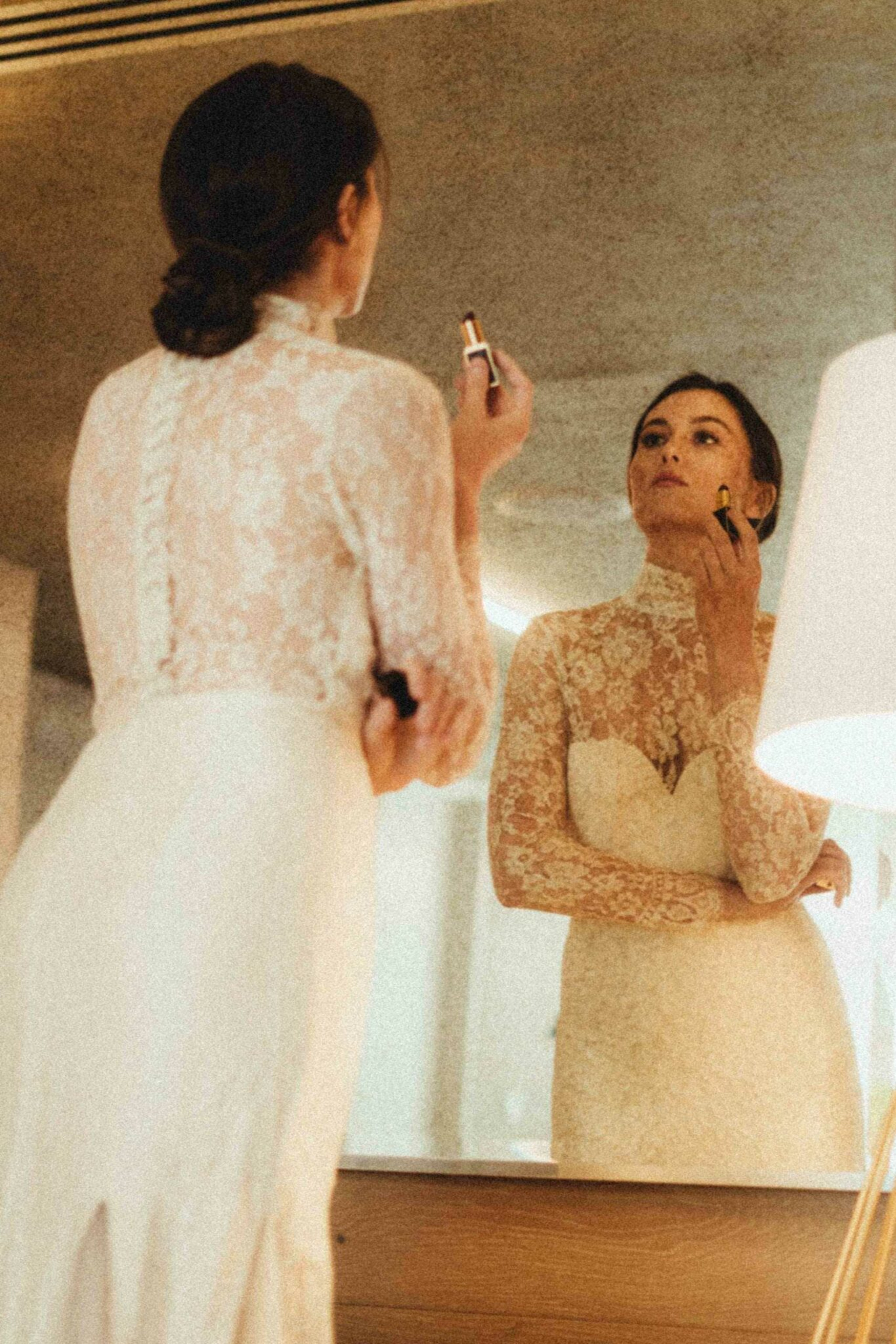 Bride in a lace wedding dress applies lipstick while looking at herself in a mirror.