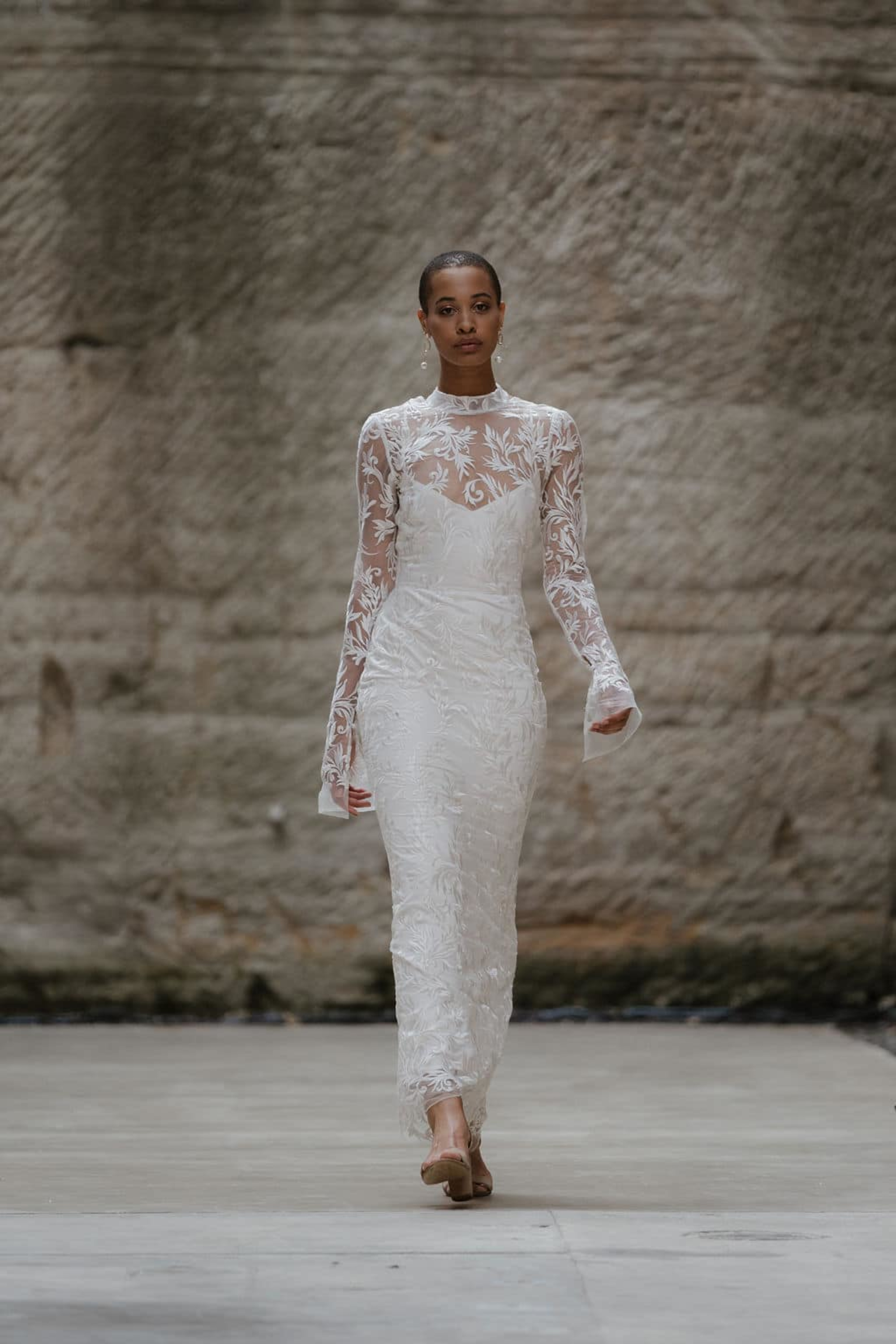 Model wearing a fitted long-sleeve lace wedding dress walking on a minimalist stone runway.